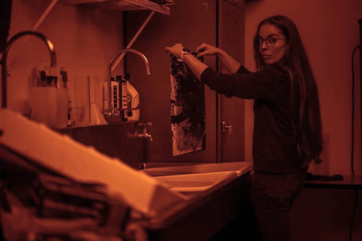 Dawn Fós standing in a photographic darkroom, washing a photographic print near a sink with pipes and shelves with bottles and supplies in the background.