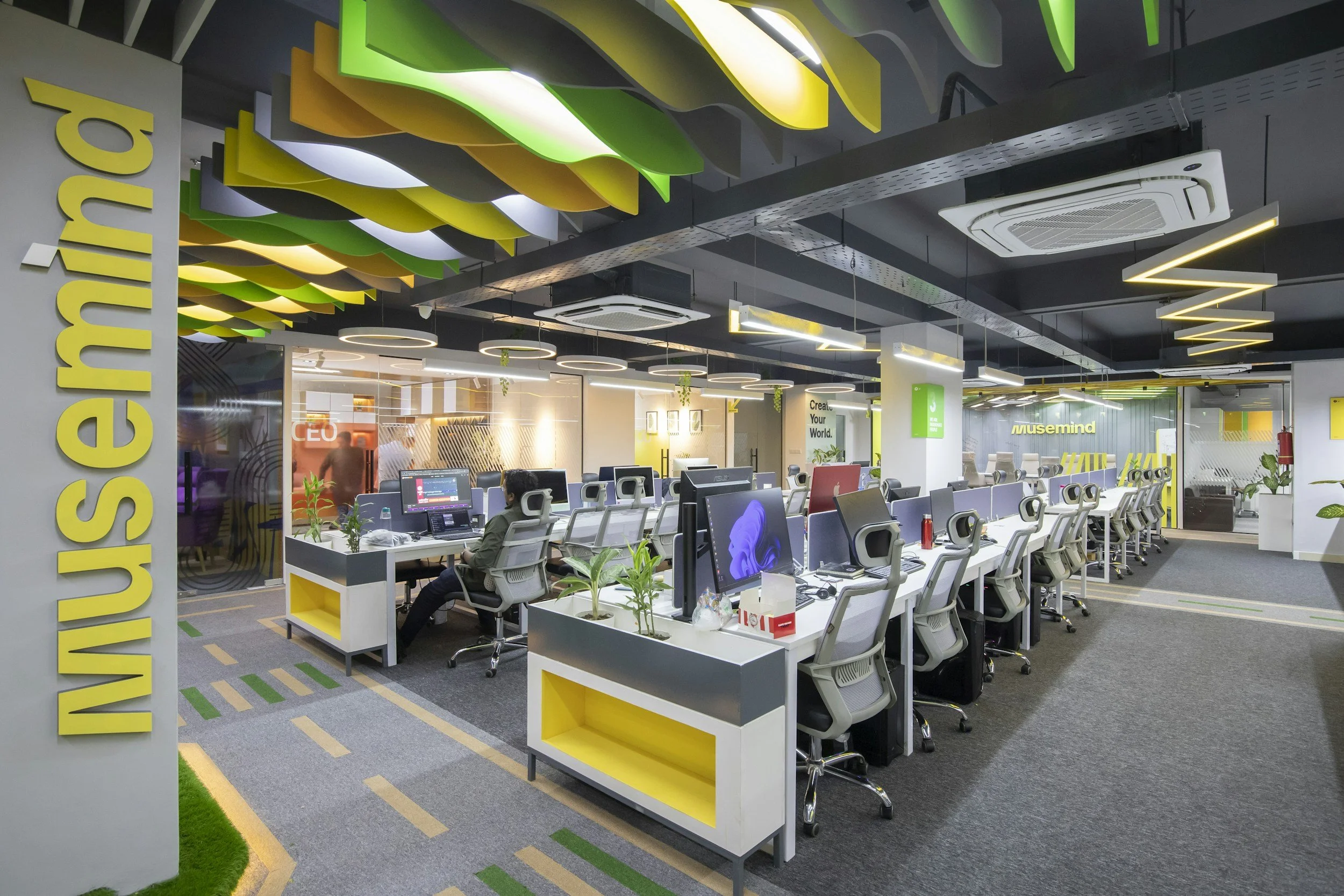Modern office workspace with rows of desks, ergonomic chairs, and multiple computer monitors. Bright lighting with decorative ceiling fixtures and green accents on the ceiling and floor. Visible branding with the word 'Musemind' and motivational signs. Employees working at their stations.
