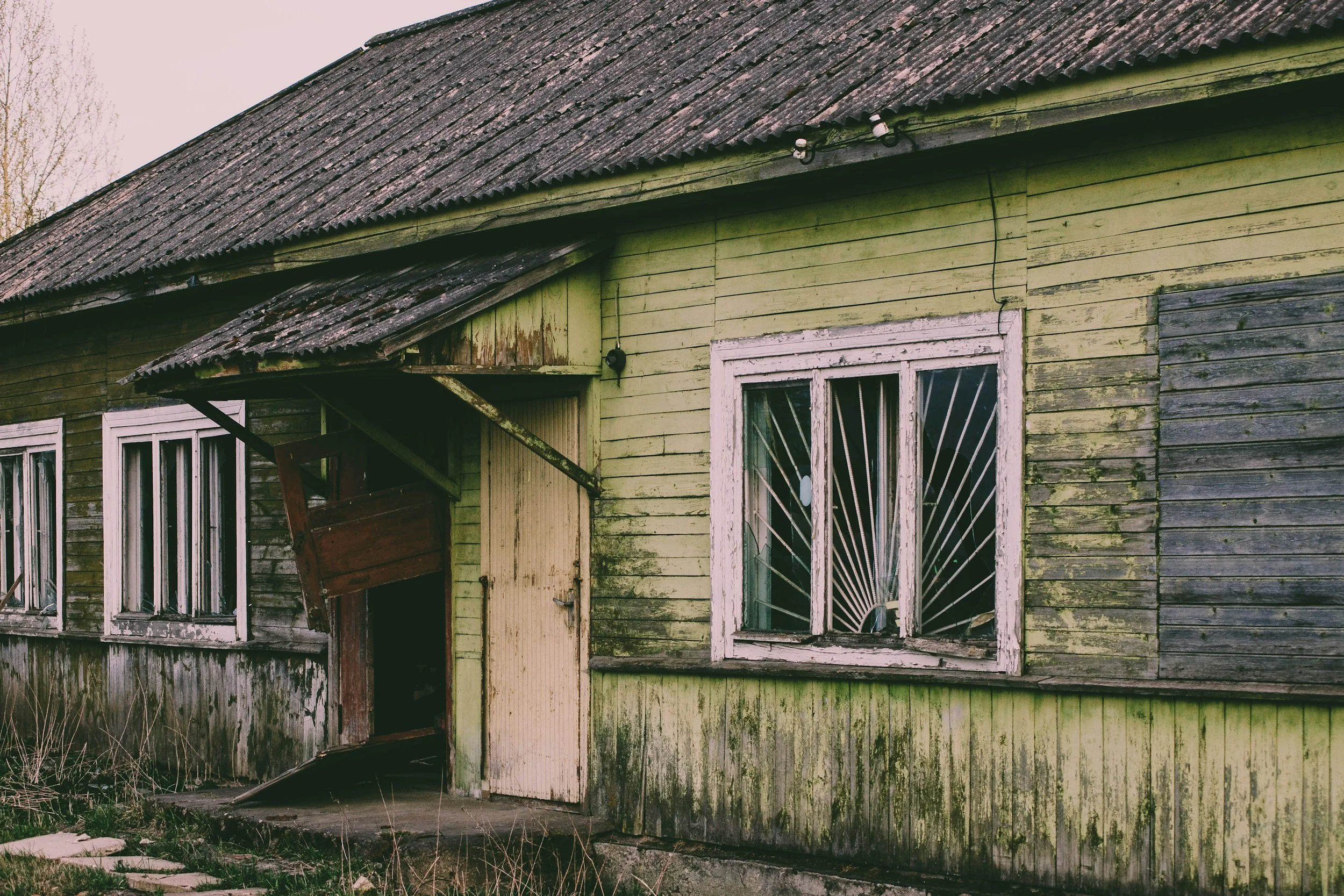 An old, weathered wooden house with peeling green paint, broken windows, and a sagging roof, with overgrown grass and bushes in front.
