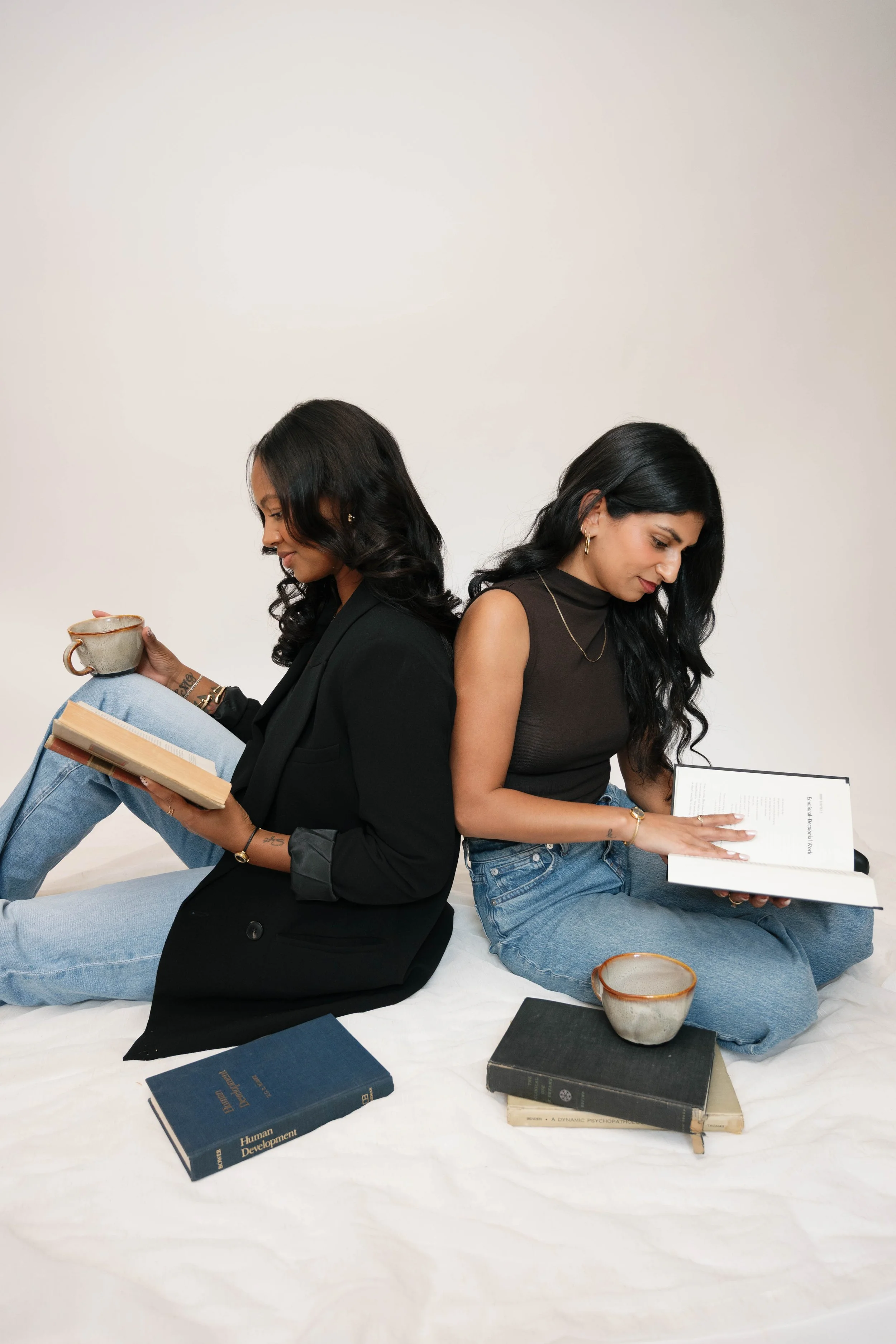 Two women sitting back-to-back, each reading a book, with books and a ceramic bowl placed around them on a white surface against a plain white background.