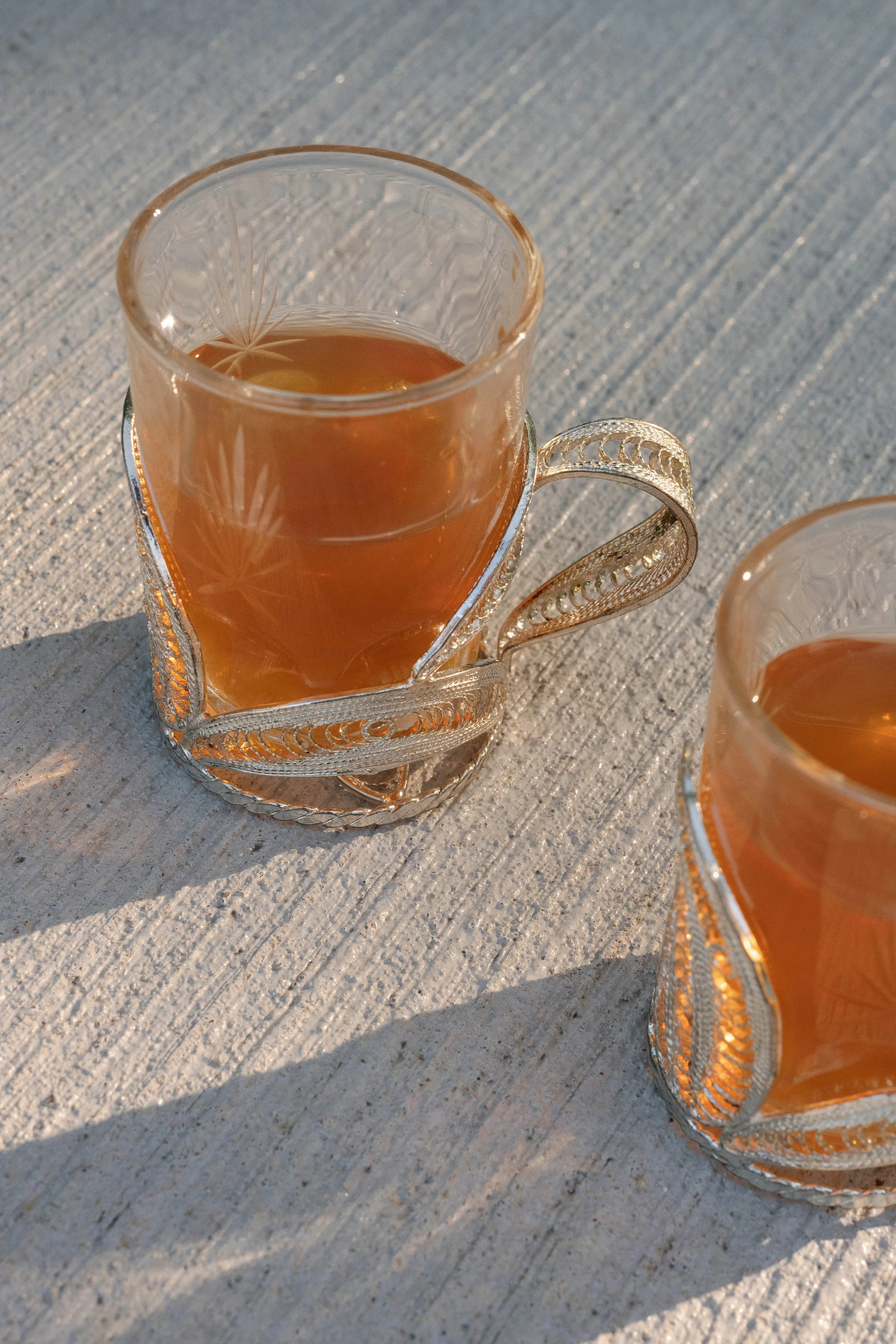 Close-up of two ornate glass cups filled with amber-colored tea, placed on a textured, light-colored wooden surface, with natural sunlight casting shadows.