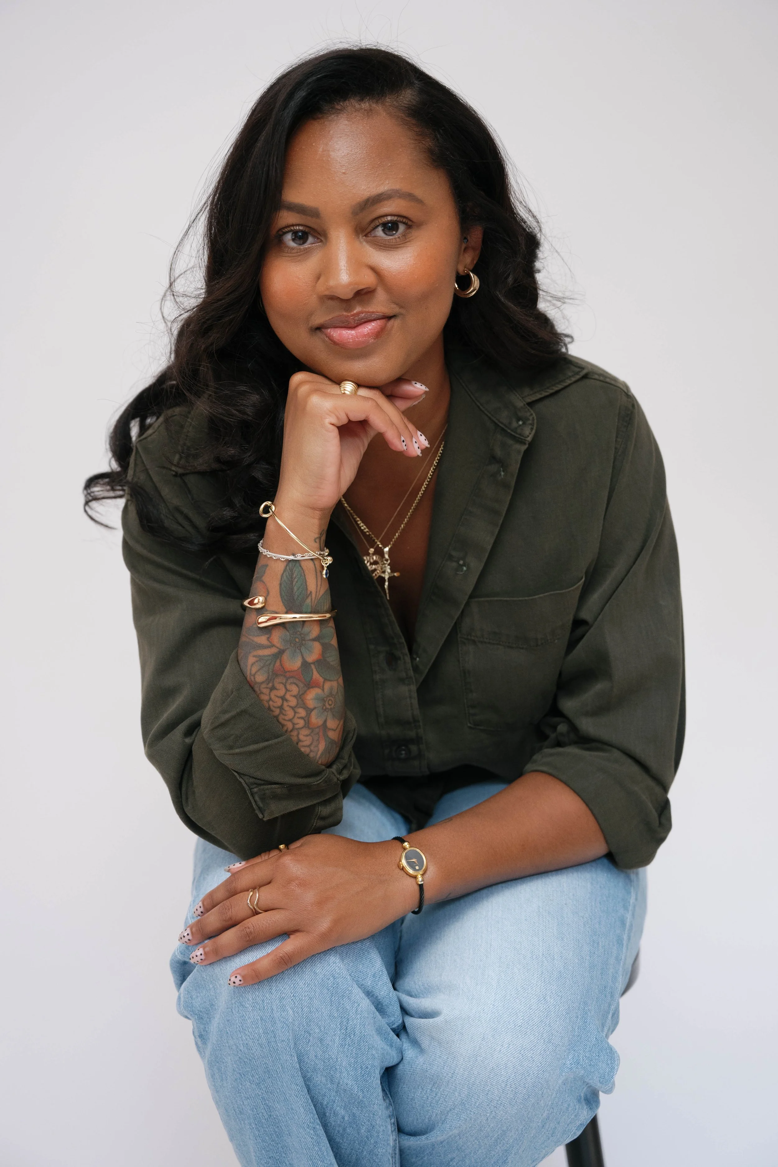 A woman with dark hair, tattoos on her arm, wearing gold jewelry and a dark green jacket, sitting against a plain white background.