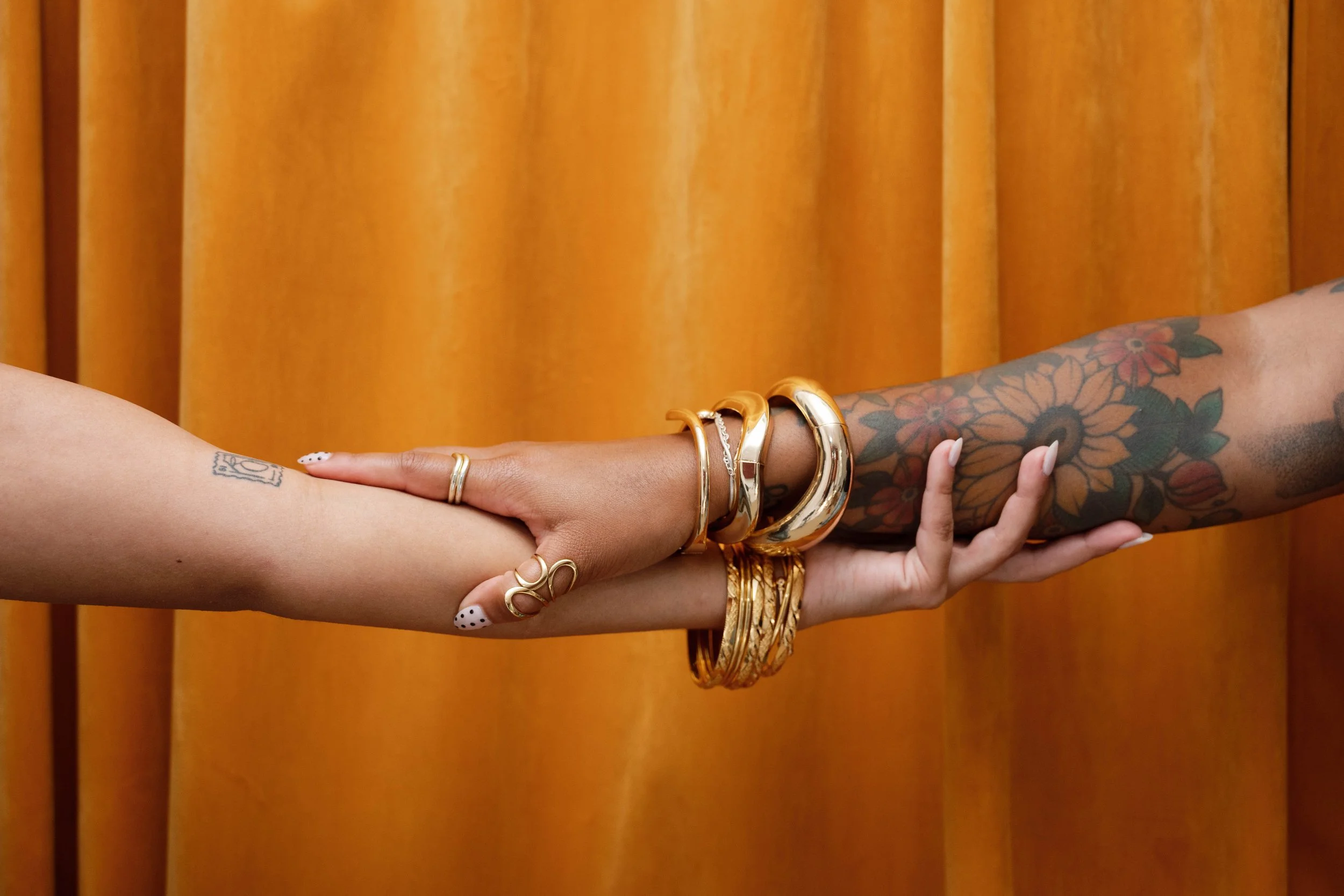 Close-up of two hands holding each other, one hand decorated with multiple gold and silver bangles and rings, along with tattoos on the arm, against a wooden background.