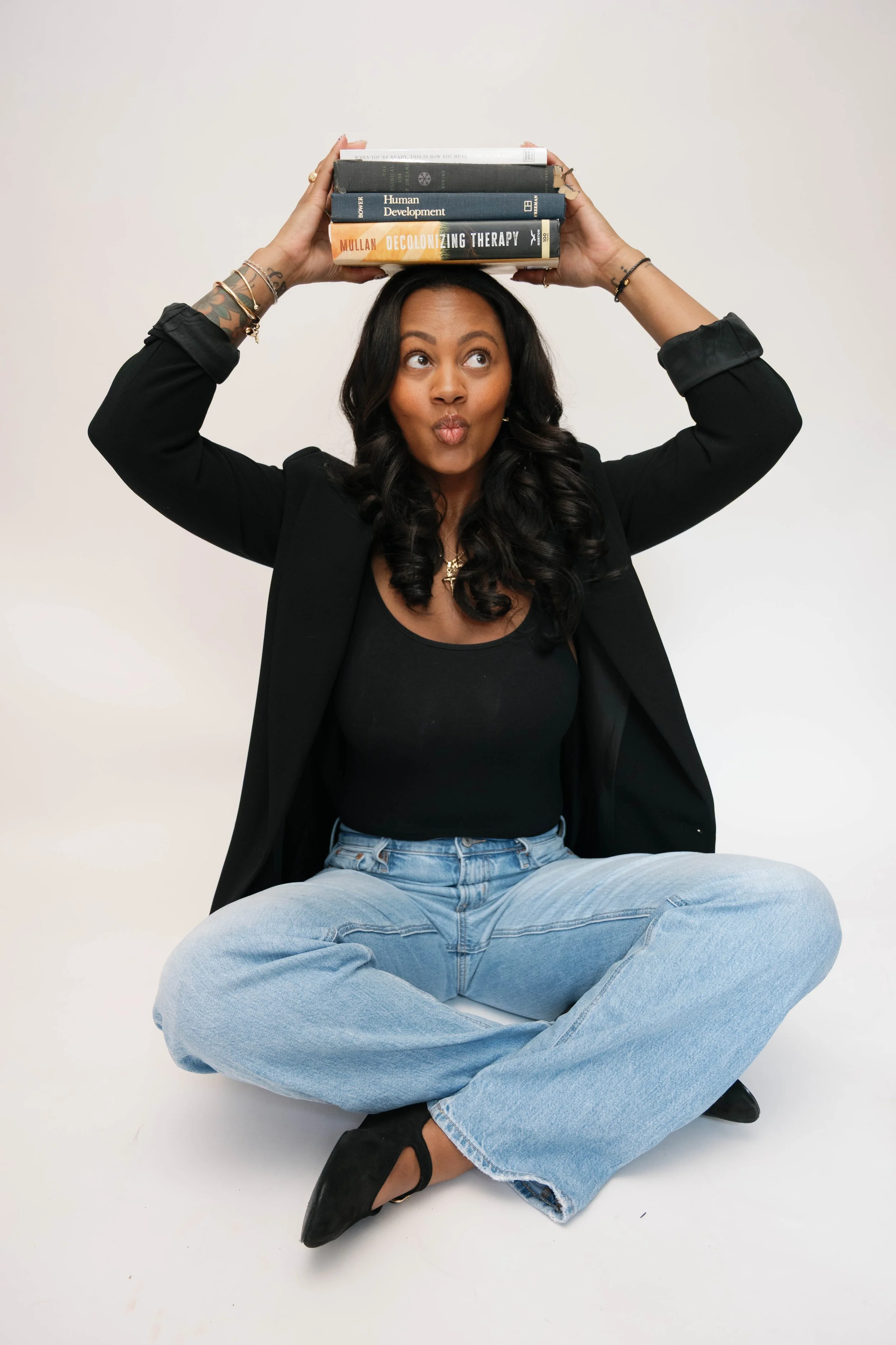 A woman with dark hair sitting cross-legged on the floor, holding four books above her head, with a playful expression and puffed cheeks.