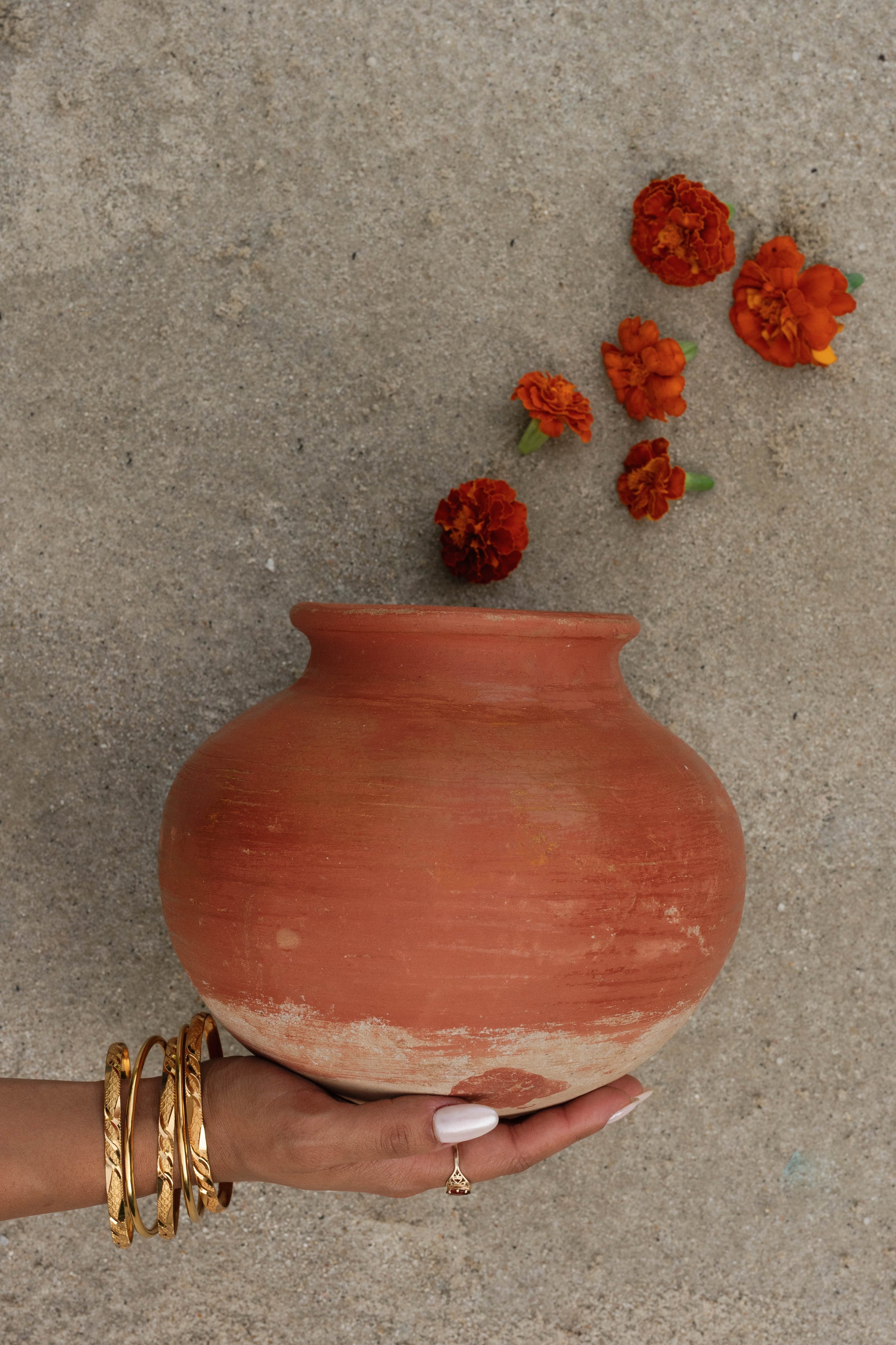 A person holding a large, round, terracotta-colored vase with orange marigold flowers spilling out of the top, set against a sandy background.