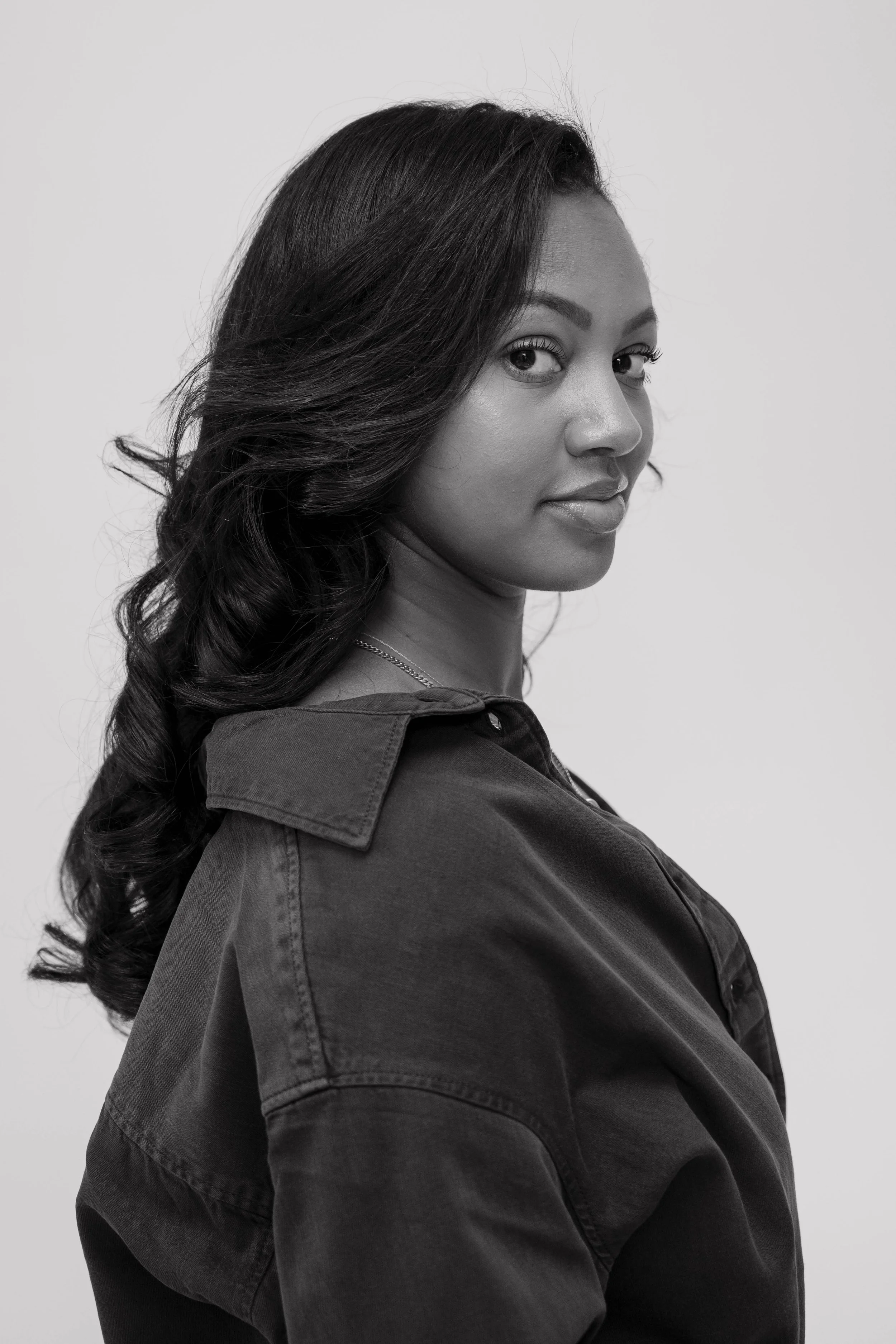 A portrait of a woman with long wavy hair, wearing a dark denim jacket, looking over her right shoulder against a plain white background.