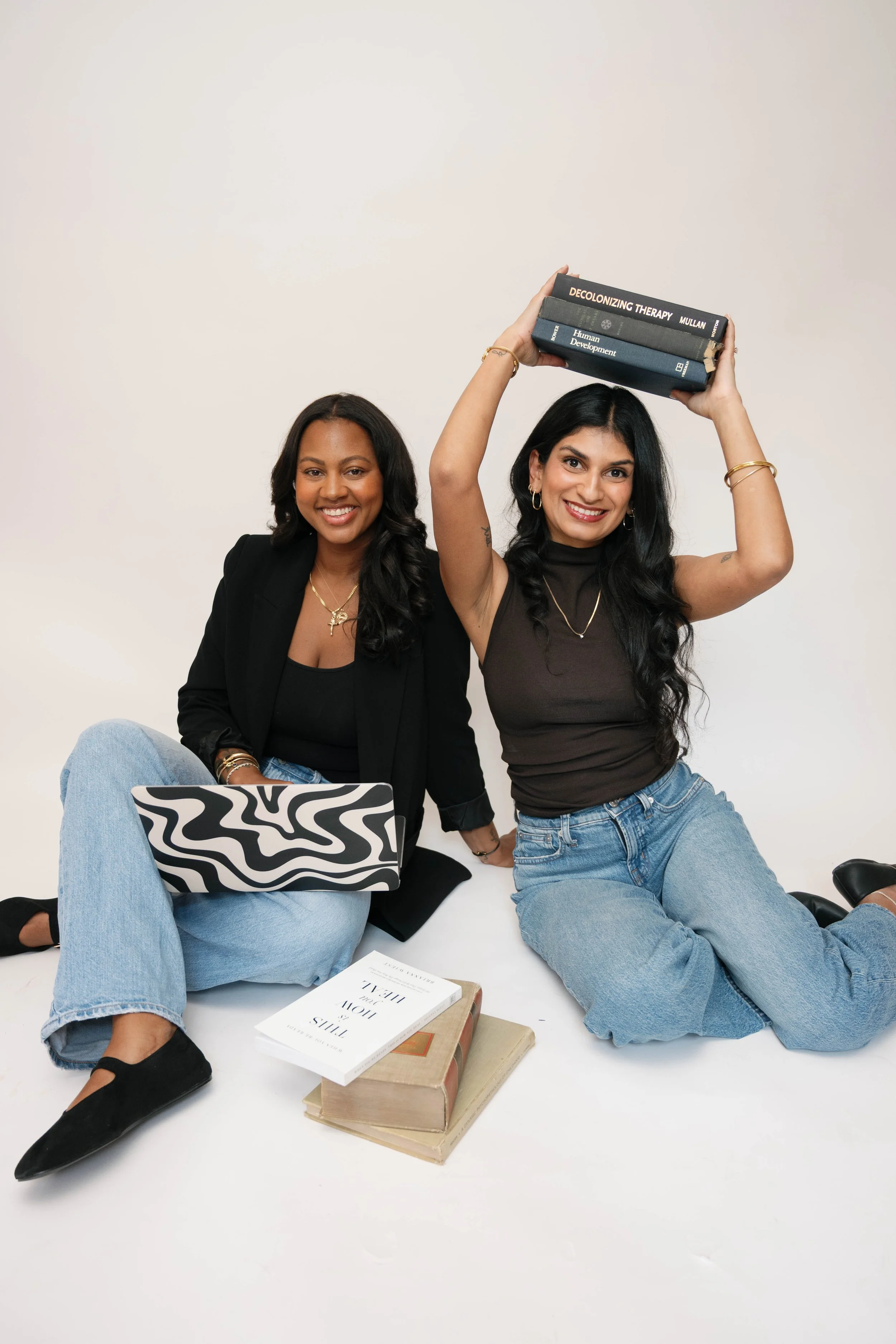 Two women sitting on the floor with a plain white background. One woman is smiling and holding a stack of books over her head. The other woman is sitting cross-legged, smiling, with a laptop and some books around her.