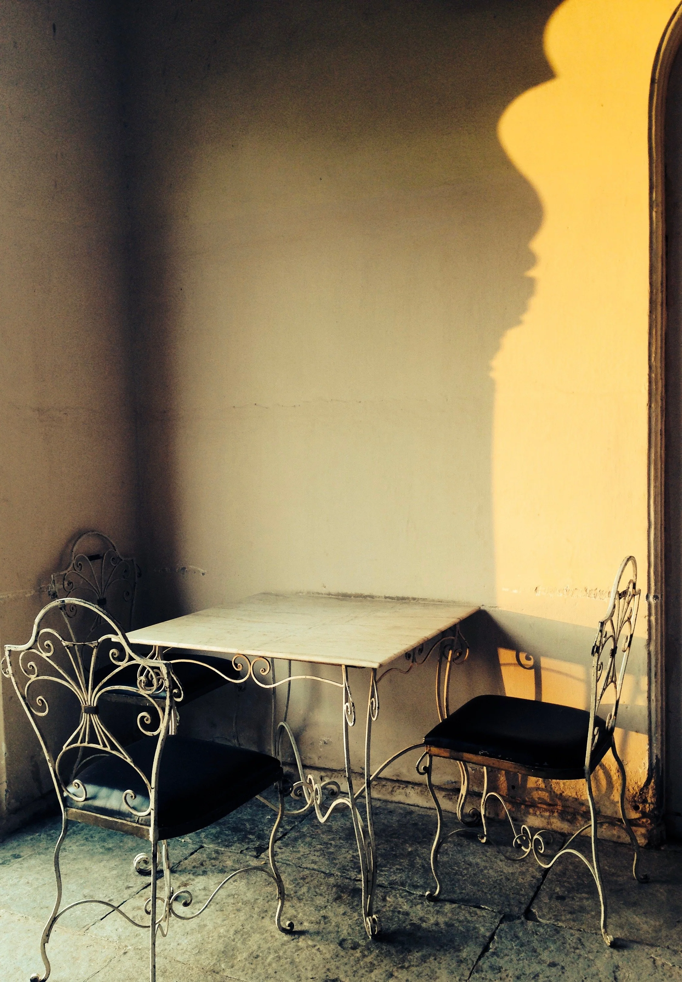 A small white metal table with a marble top and four matching black cushioned metal chairs with decorative backrests, set against a beige wall with shadows cast by the chairs and table, on a concrete floor.
