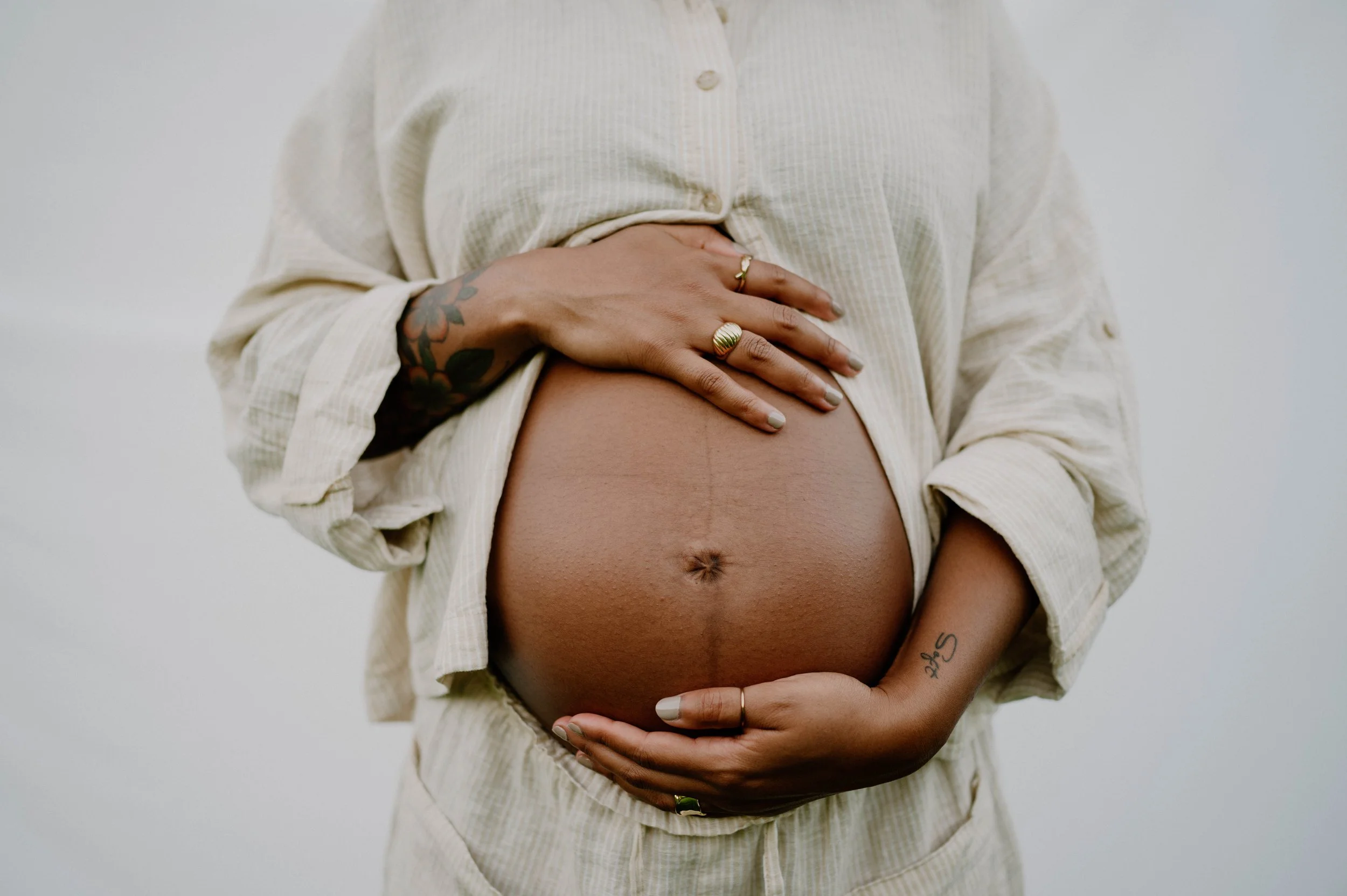 A pregnant person wearing a light-colored, textured button-up shirt, with hands resting on their bare belly. The person has multiple rings on their fingers, visible tattoos on their left hand, and a light skin tone.
