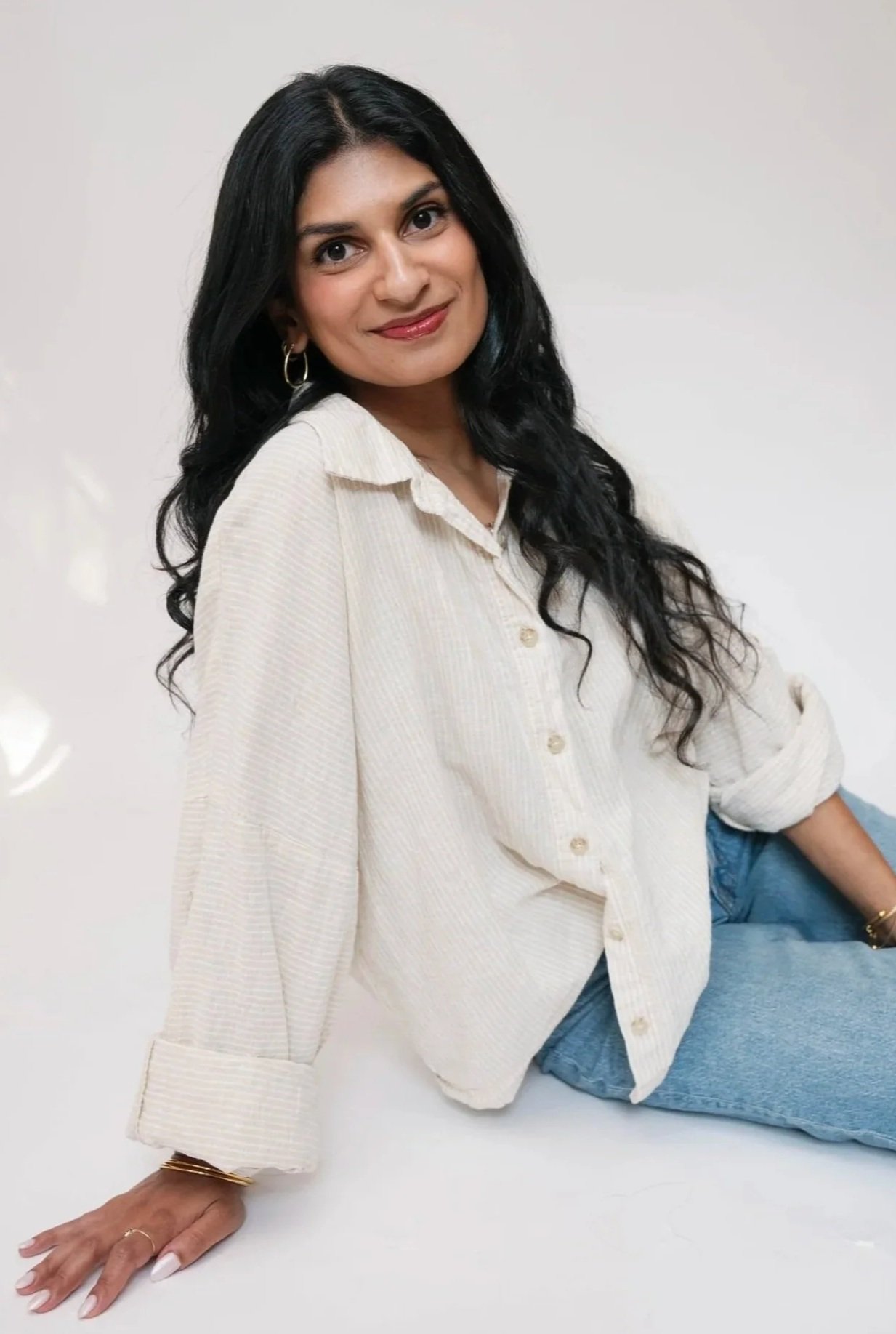 A woman with long black curly hair wearing a cream-colored oversized button-up shirt and blue jeans, sitting and leaning on her left hand, against a plain white background.