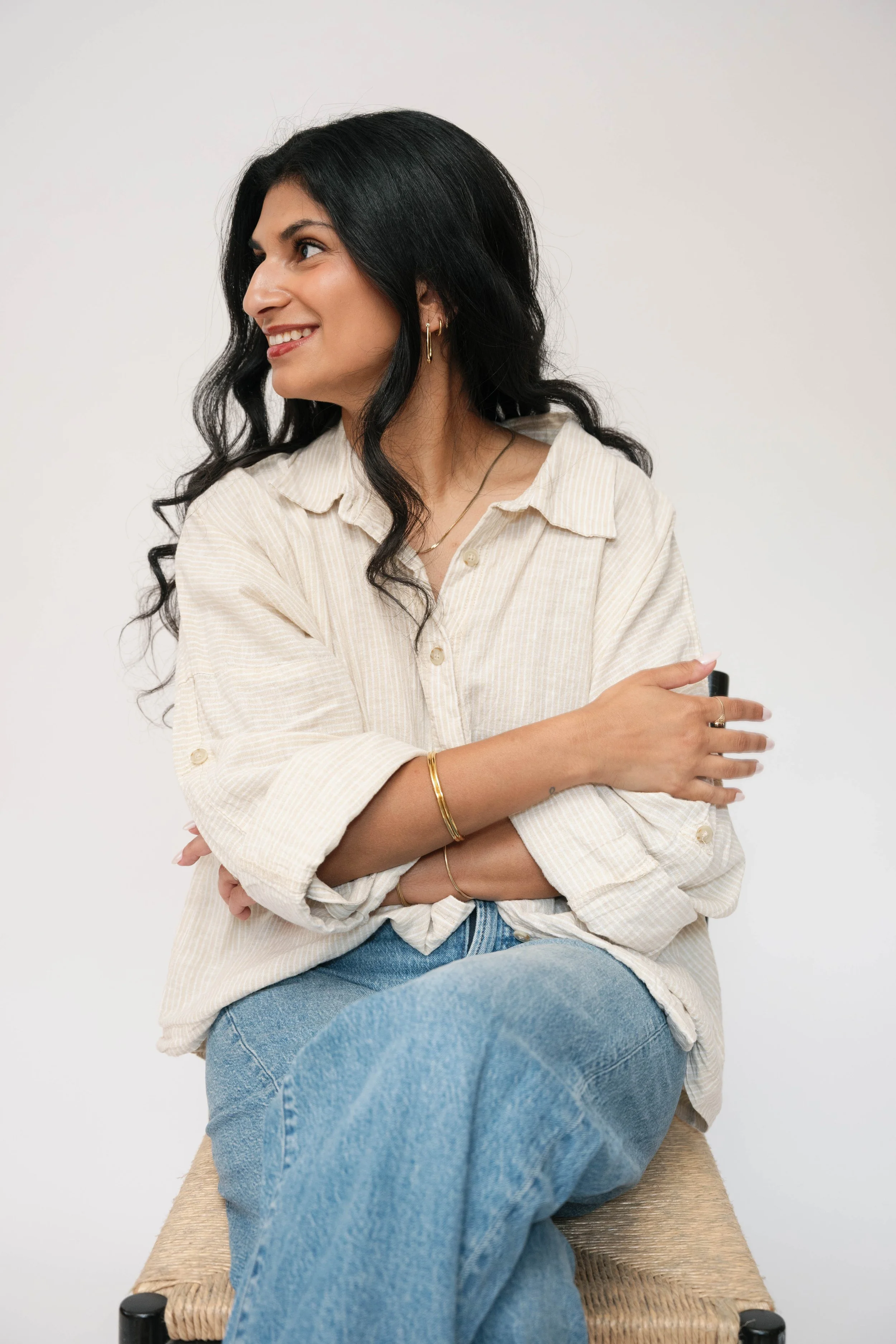 A woman with long black hair, wearing a beige striped button-up shirt and blue jeans, sitting cross-legged on a woven chair with arms crossed, smiling and looking to her left against a plain white background.
