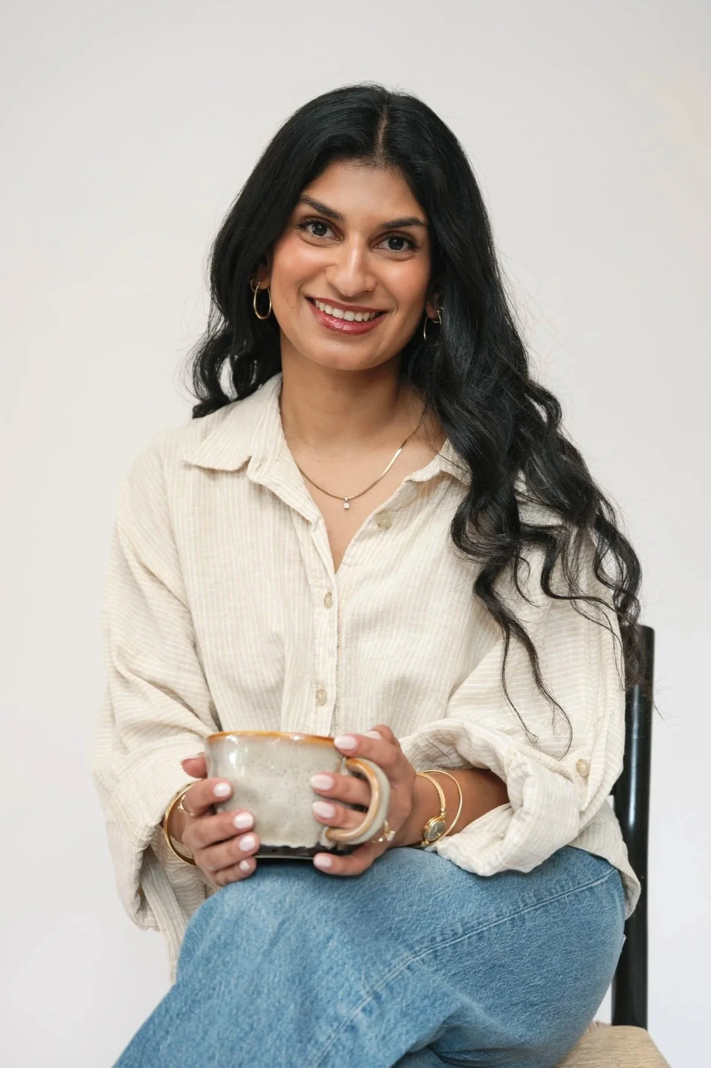 A woman with long dark hair, wearing a beige button-up shirt, silver hoop earrings, and a gold necklace, sitting on a beige chair and holding a ceramic mug, smiling against a plain white background.