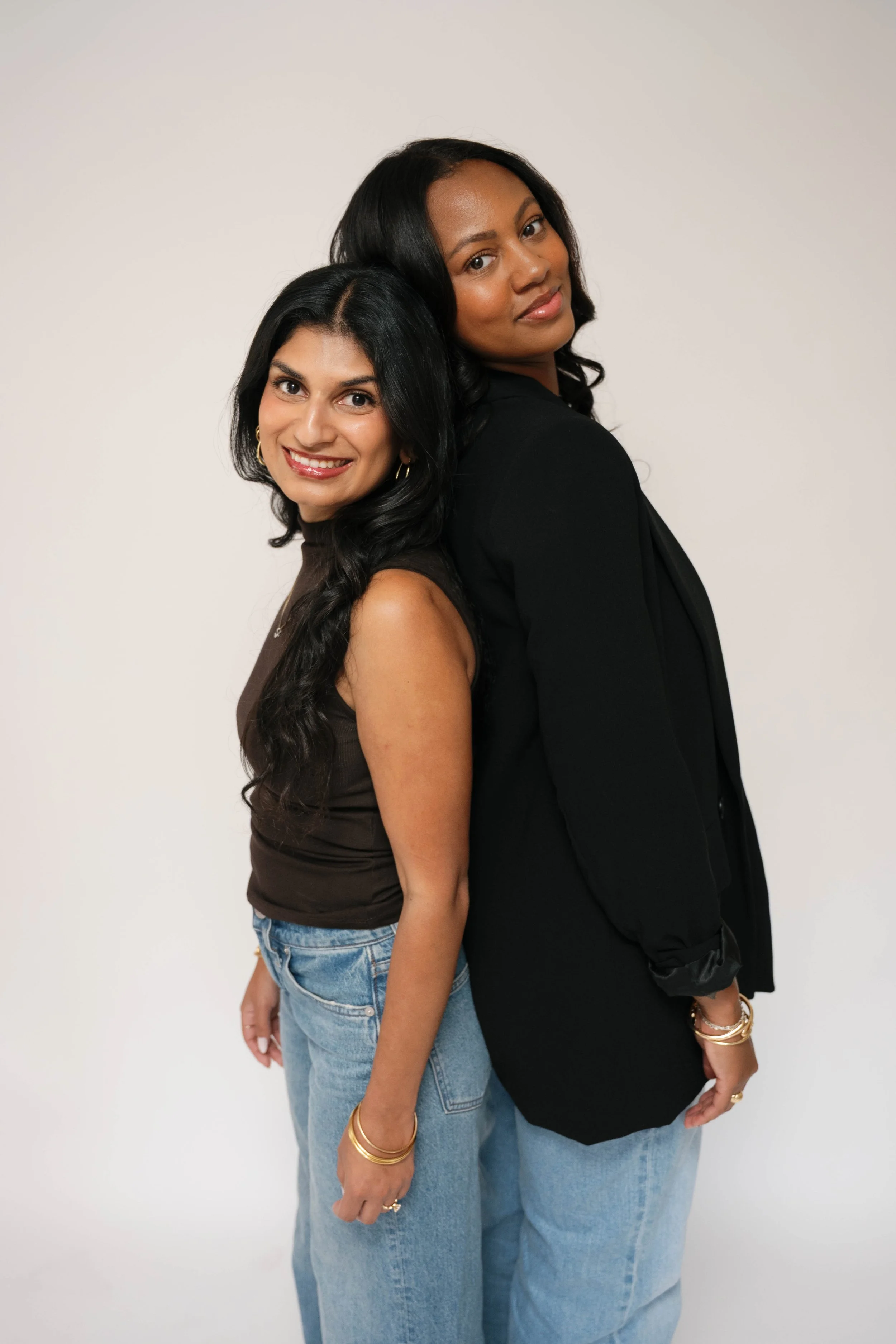 Two women standing back to back, smiling, with a plain light-colored background.