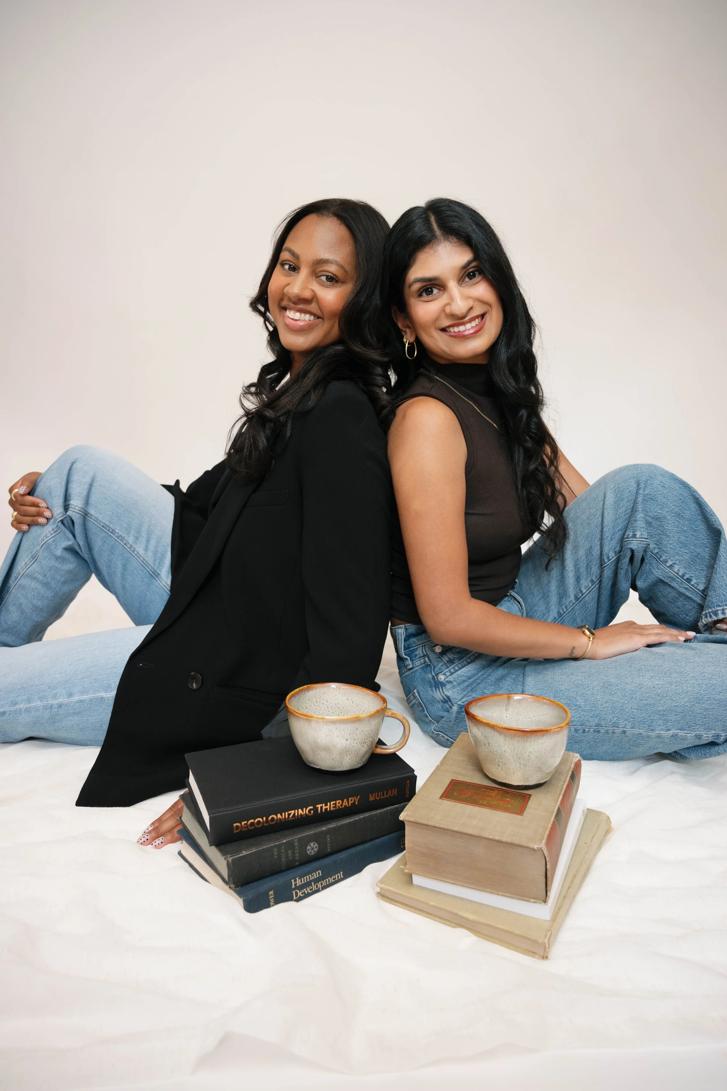 Two smiling women sitting back-to-back on a white sheet, surrounded by books and cups, with one woman wearing a black blazer and the other in a sleeveless black top and jeans.