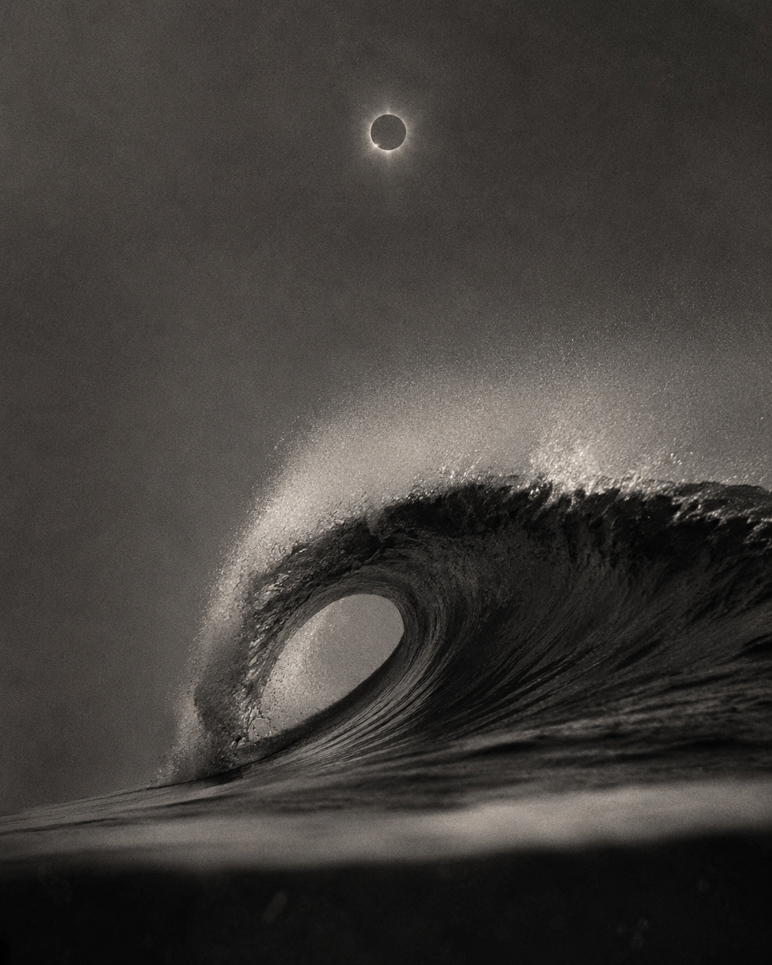 A black and white photograph of an ocean wave curling with a visible hollow inside, and the moon shining in the dark sky above.