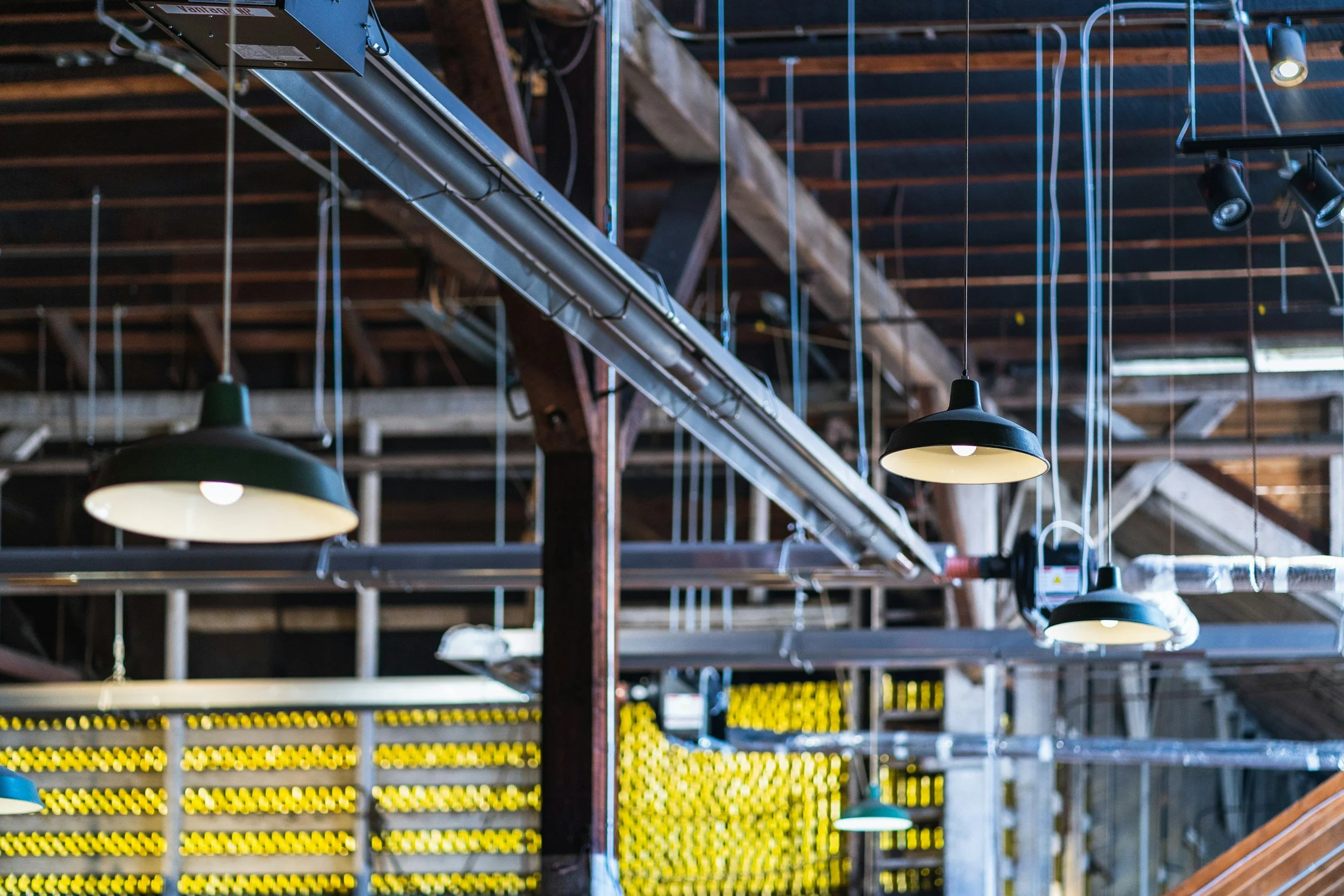 Industrial ceiling with hanging black pendant lights, exposed wooden beams, and a metal duct work. There are yellow and black safety markings on the wall below.
