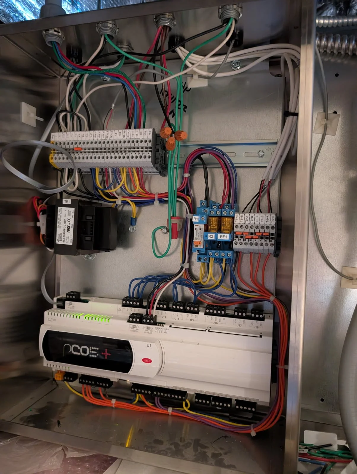A close-up of an electrical control panel with various wired components, switches, relays, and circuit breakers mounted on a metallic frame.