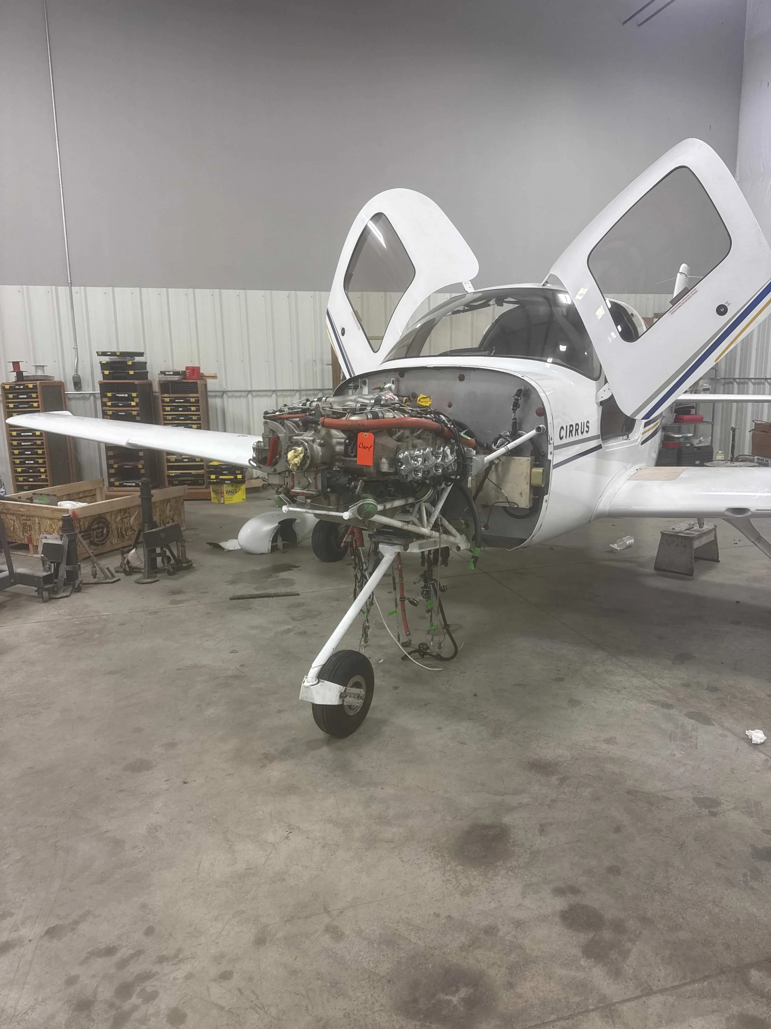 A partially assembled aircraft with its engine exposed is inside a workshop. The aircraft's wings are attached, but the fuselage is open at the front. The workshop has tools and storage shelves nearby. Cirrus SR20.
