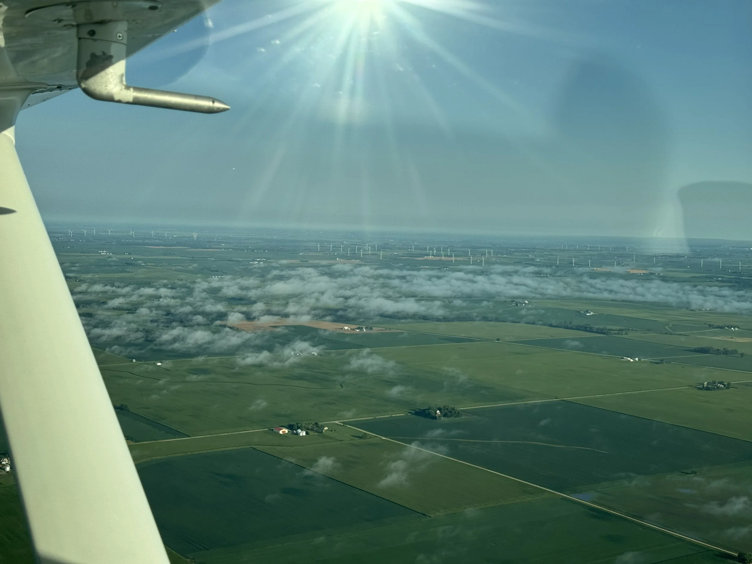 Aerial view of farmland and fields with wind turbines in the distance, seen from an airplane in flight during daytime.