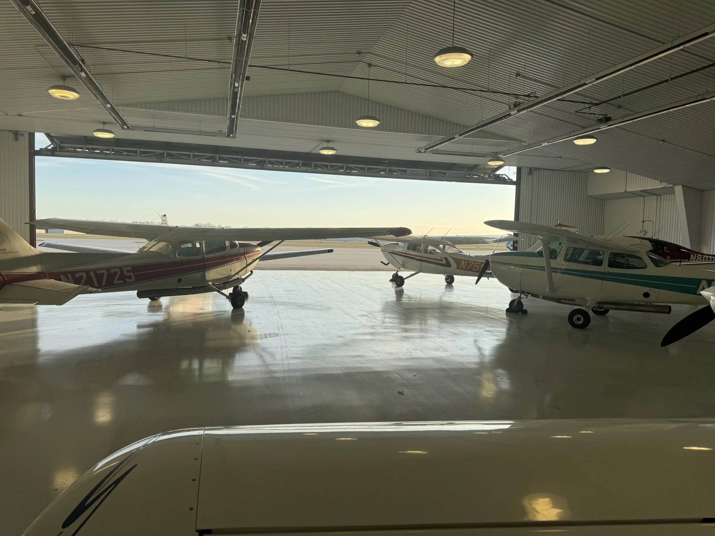 Inside a hangar with a corrugated metal ceiling, four small airplanes are parked near the open hangar door, with a view of the runway and sky outside. Fleet Support.