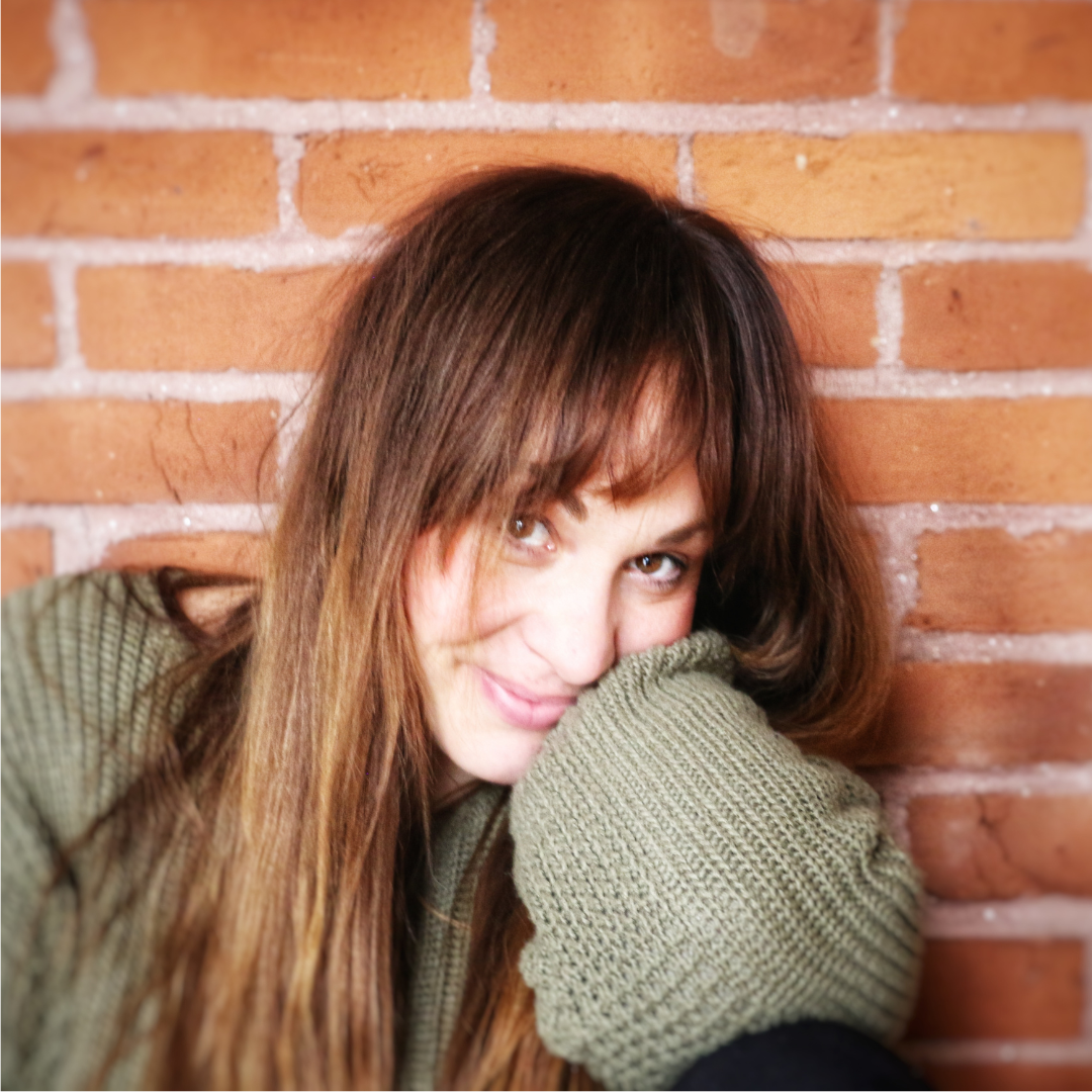 Jessica Proulx long brown hair and light eyes leaning against a brick wall, smiling with her face partially hidden behind her arm.