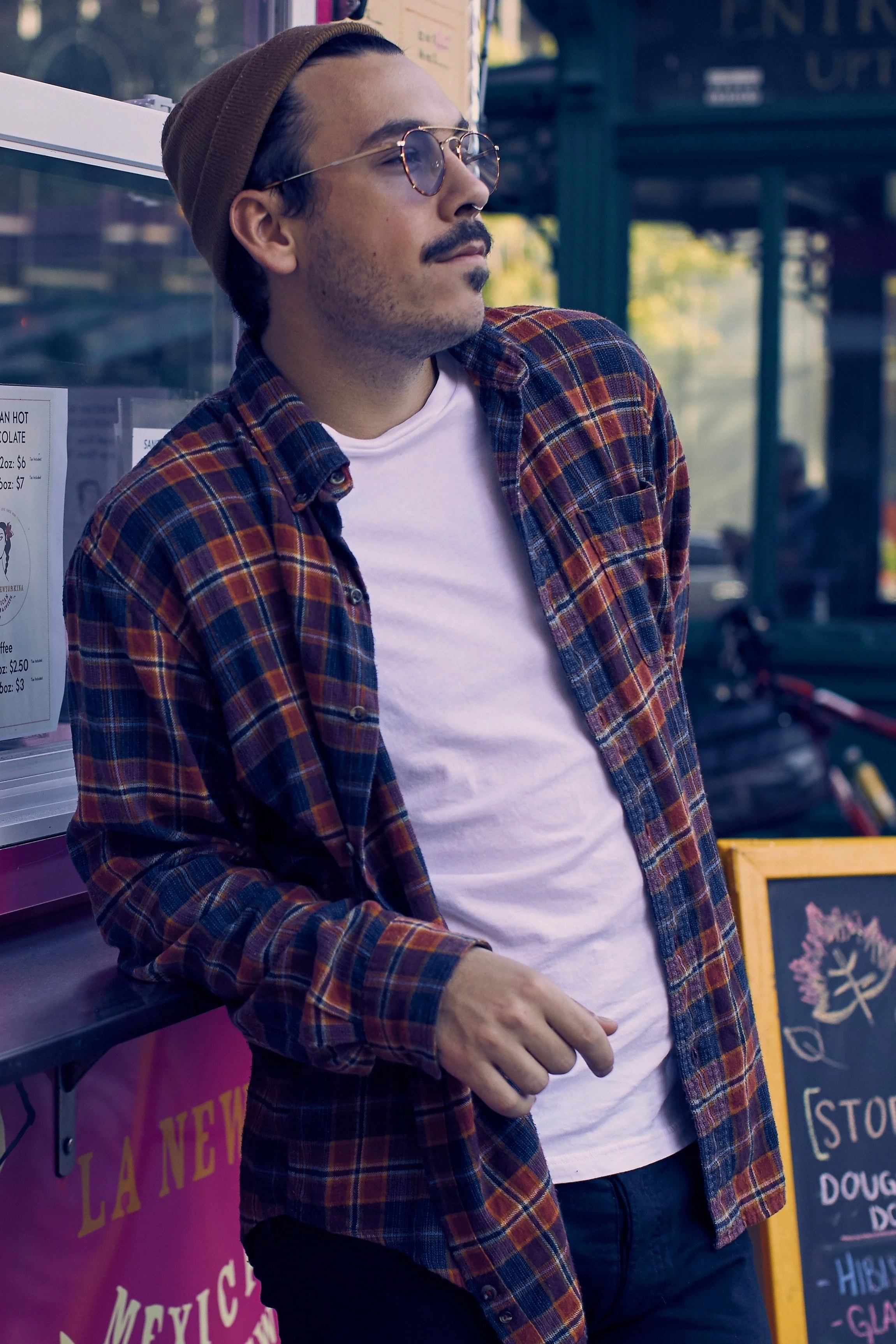 Billy Gaffney with sunglasses, a brown beanie, a mustache, and beard, wearing a plaid shirt over a white t-shirt, leaning against a pink wall with outdoor menu signs in the background.
