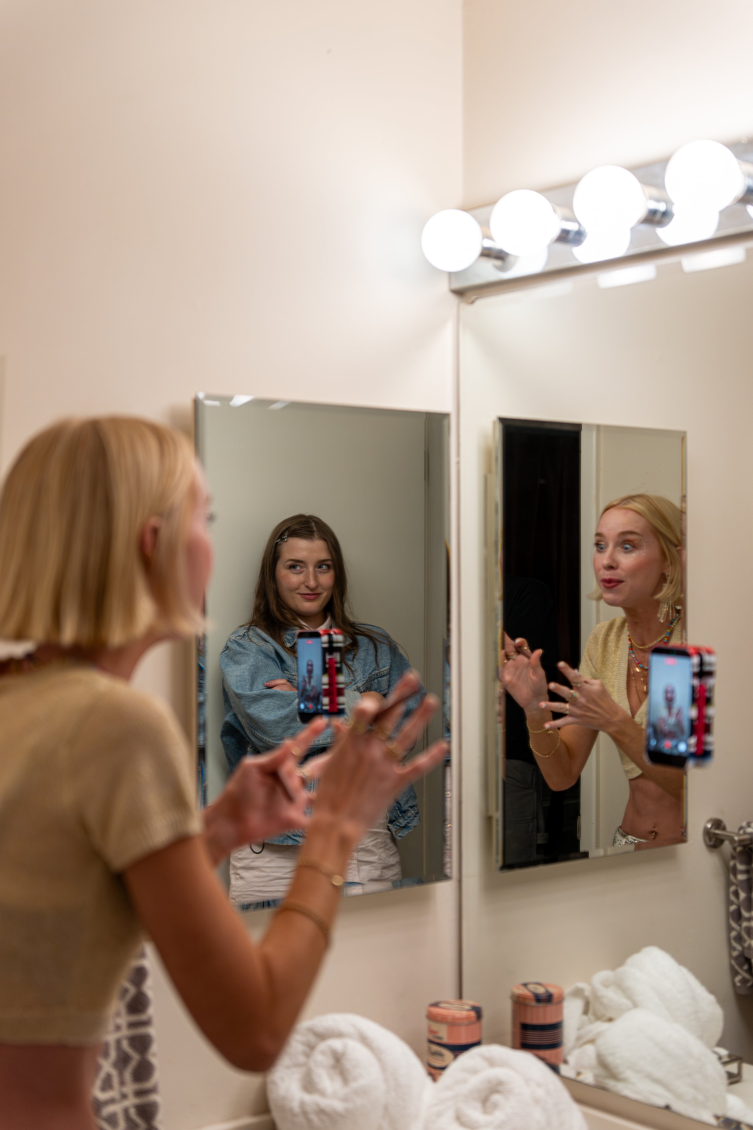 A woman with blonde hair taking a selfie in a mirror, with another woman with brown hair also in the mirror, and a woman with blonde hair talking, all in a bathroom with a row of four round vanity lights, shelves with rolled white towels, and various skincare products.