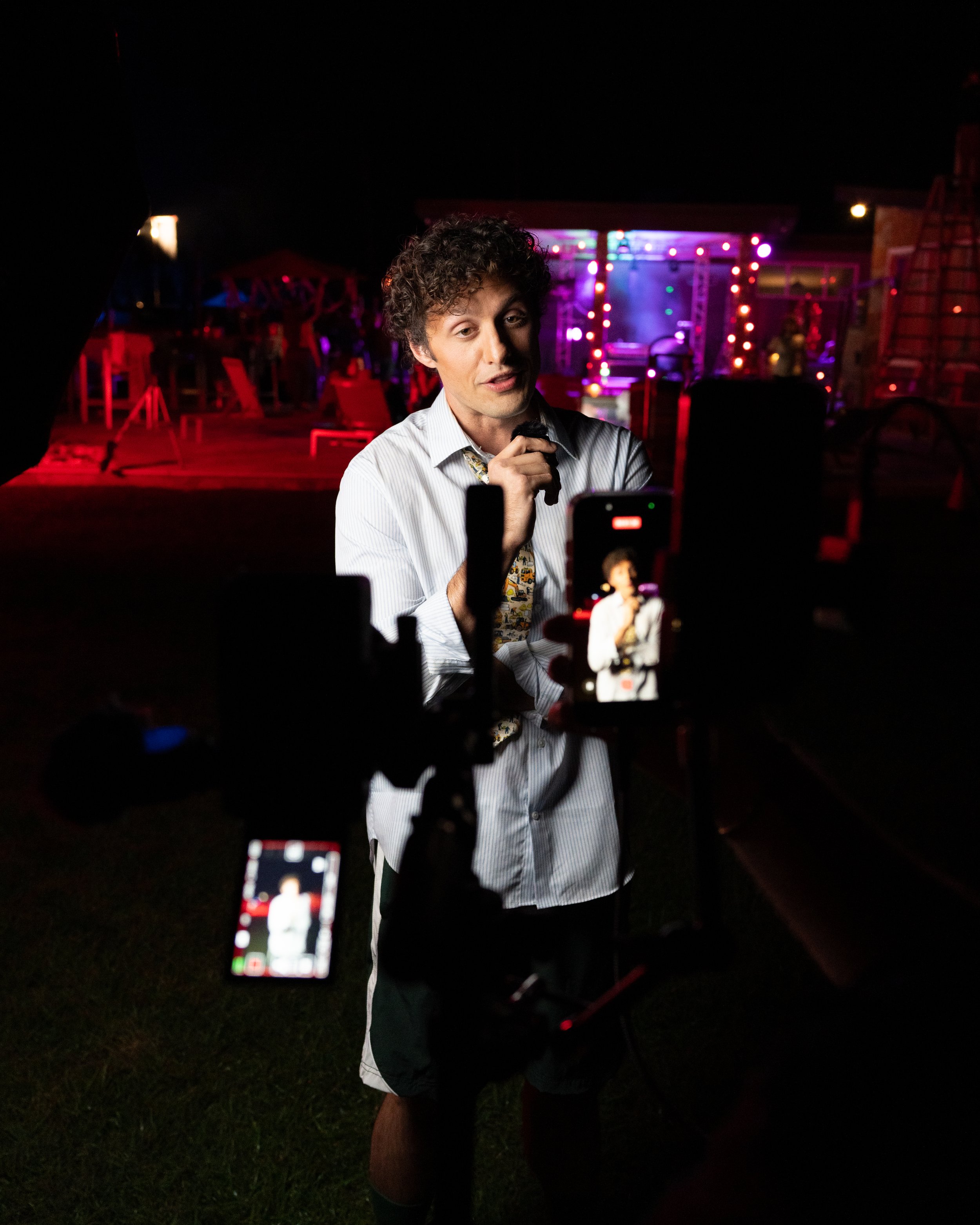 A man with curly hair wearing a striped shirt and patterned tie is being filmed or photographed at night, with colorful lights and an outdoor stage in the background. Multiple people are capturing the scene with their phones.
