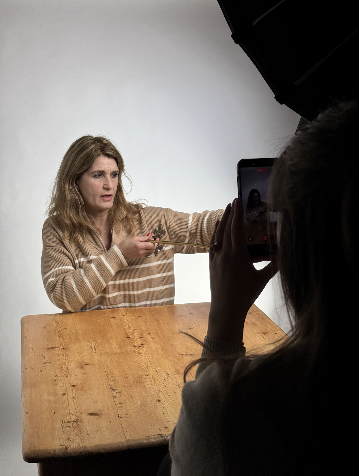 A woman is sitting at a wooden table, holding a metal object, while a person takes a photo or record video of her with a smartphone. The setting appears to be a studio with a plain background.