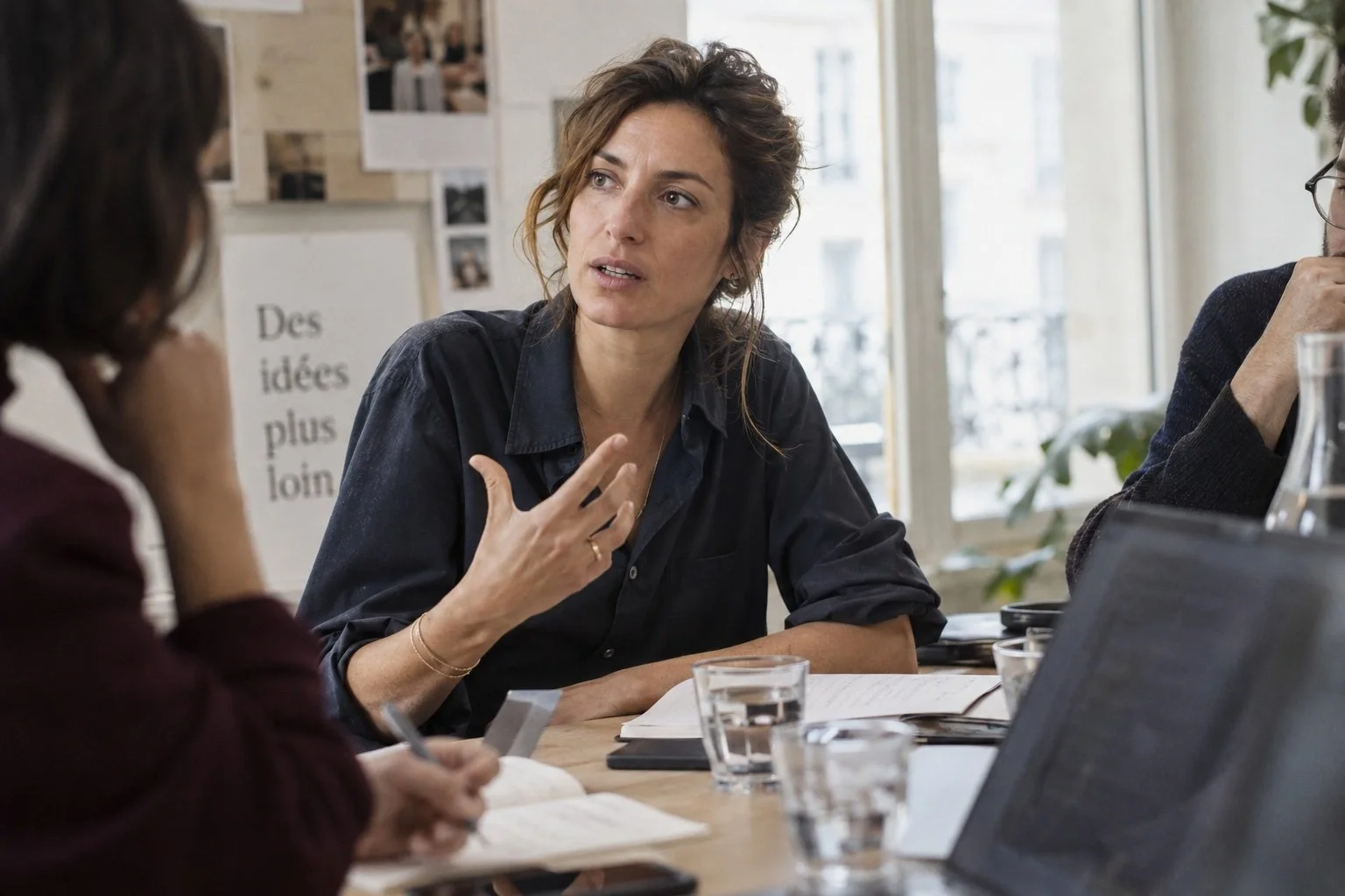 Une femme discute lors d'une réunion avec deux autres personnes dans une salle lumineuse, avec des fenêtres visibles à l'arrière-plan.