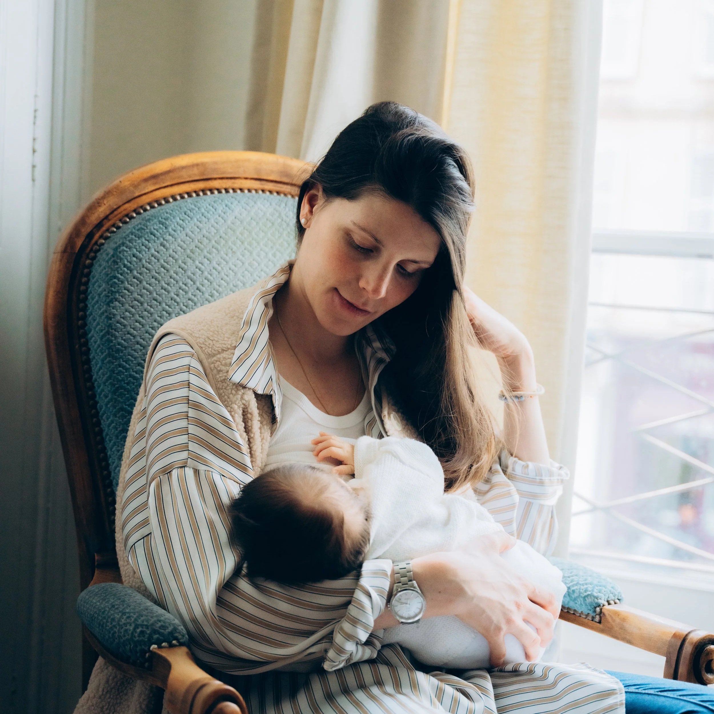 Une jeune femme aux cheveux bruns, portant une chemise rayée et une montre, est assise sur une chaise en bois avec un revêtement en tissu vert. Elle tient un bébé blanc dans ses bras. La scène se déroule près d'une grande fenêtre avec des rideaux jaunes, laissant entrer beaucoup de lumière naturelle.
