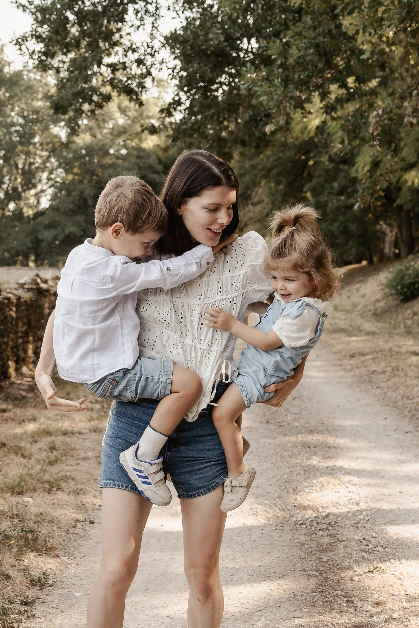 Une femme portant deux enfants dans un parc, entourée d'arbres, lors d'une journée ensoleillée.