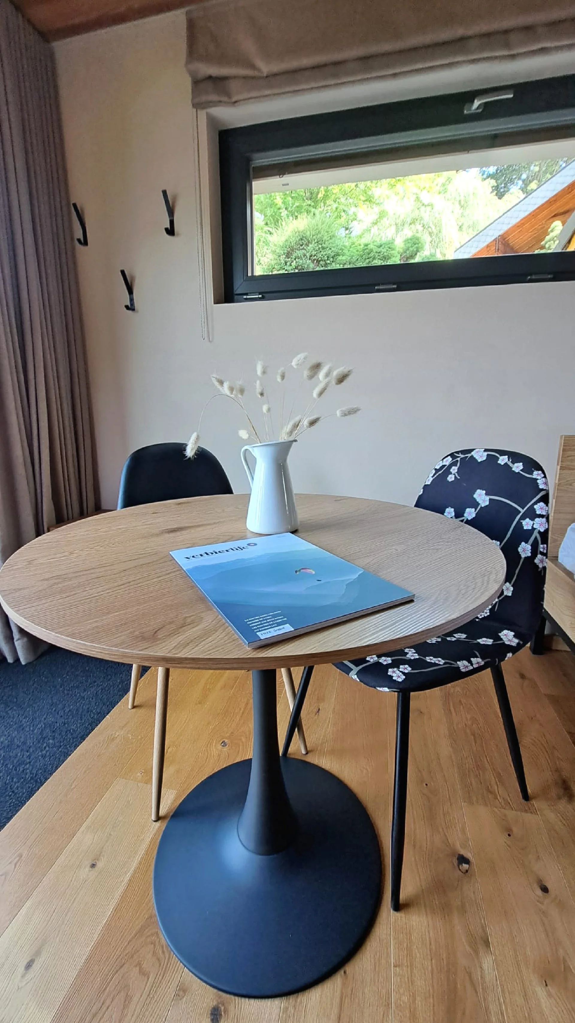 Une table en bois avec une vase blanche contenant des herbes séchées, un magazine dessus, et deux chaises, une noire et une avec un tissu fleuri, dans une salle avec une fenêtre ouverte et un mur beige.