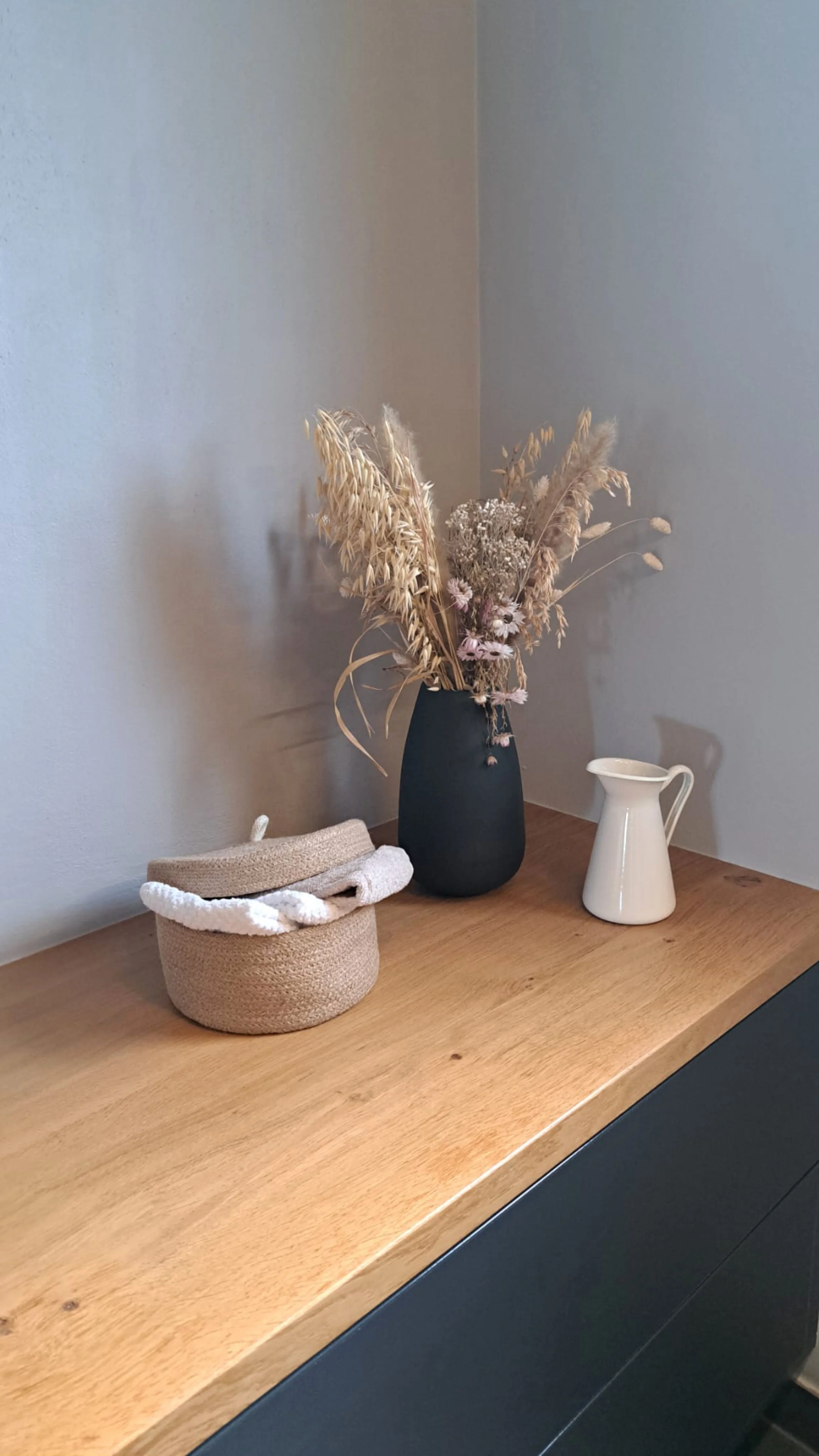 Vase noir avec des fleurs séchées, panier en osier avec un chapeau posé dessus, petite cruche blanche sur une surface en bois.