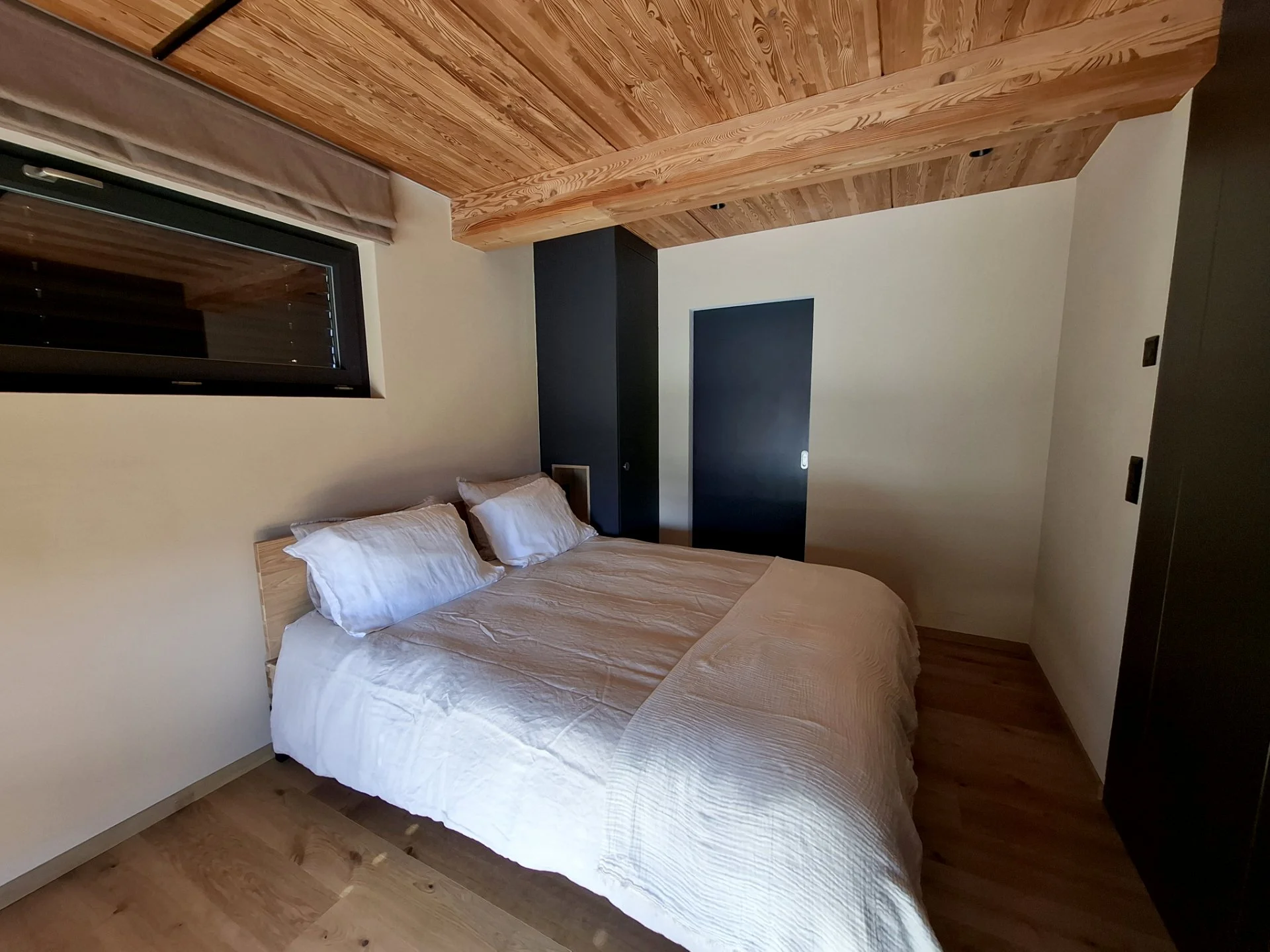 Chambre avec un lit double avec draps blancs, murs clairs, fenêtre avec voilage et mobilier en bois sombre.