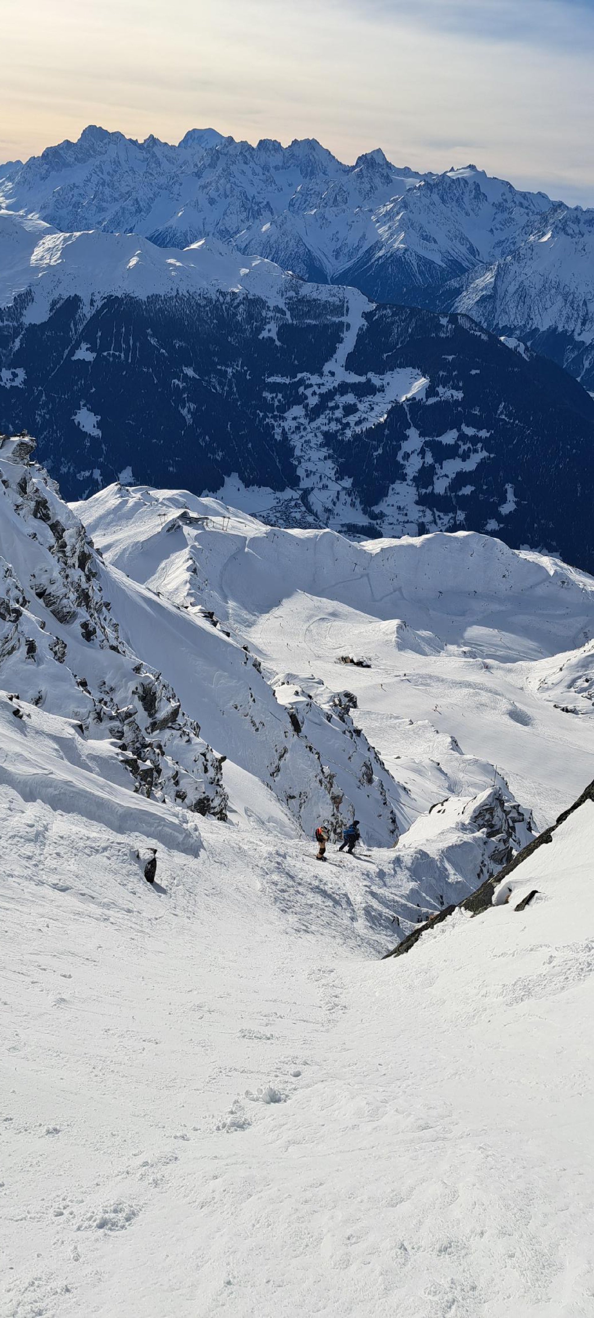Montagnes enneigées avec deux skieurs en bas, vue panoramique de sommets alpins, ciel nuageux.