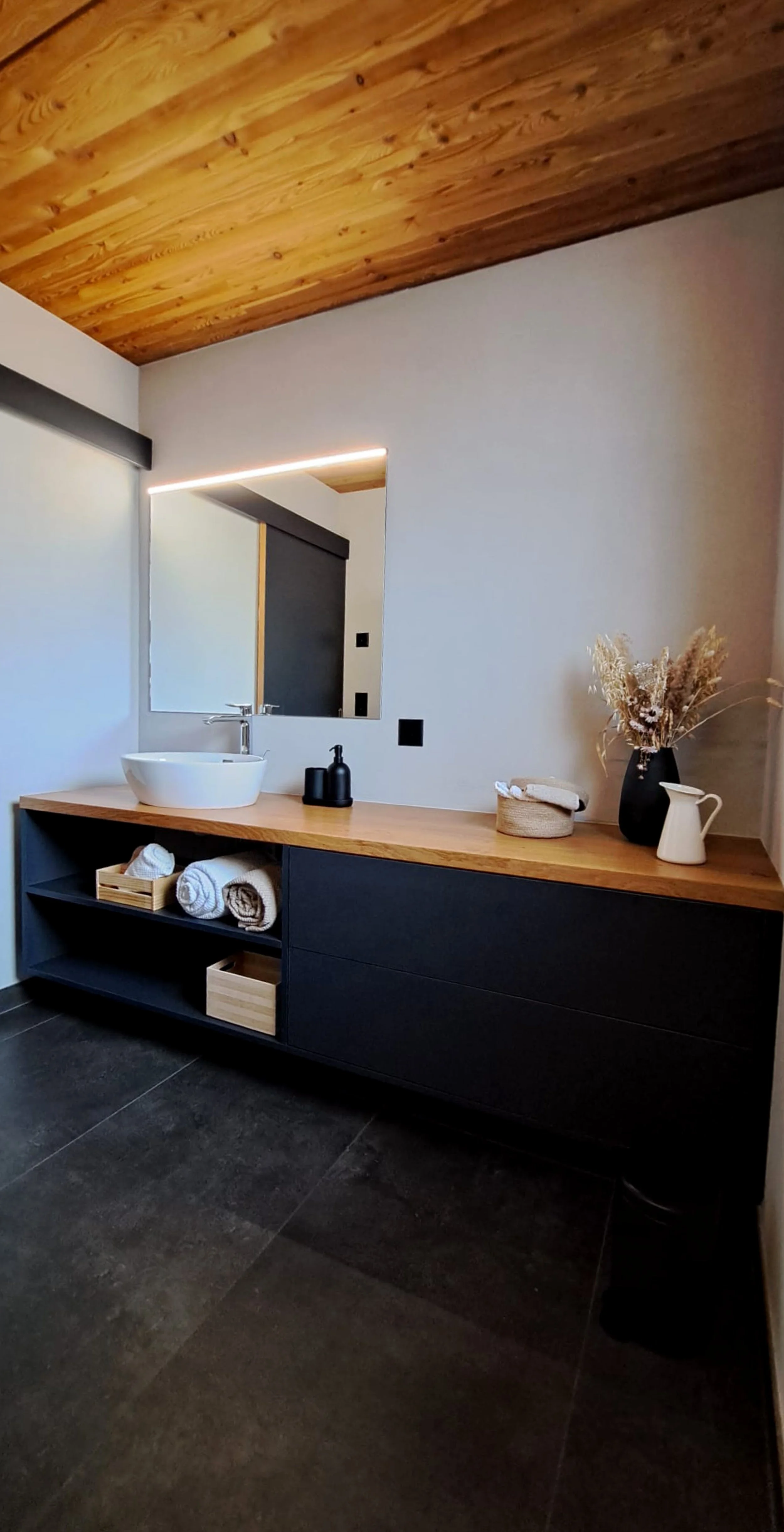 Un meuble de salle de bain noir avec un plan de travail en bois clair. Sur le plan de travail, il y a un lavabo blanc rond, un distributeur noir, une petite panière en rotin avec une serviette blanche, une vase noire avec des fleurs séchées, une cruche blanche et une petite cruche blanche. Dessous, il y a des étagères contenant des serviettes roulées et des boîtes en bois.