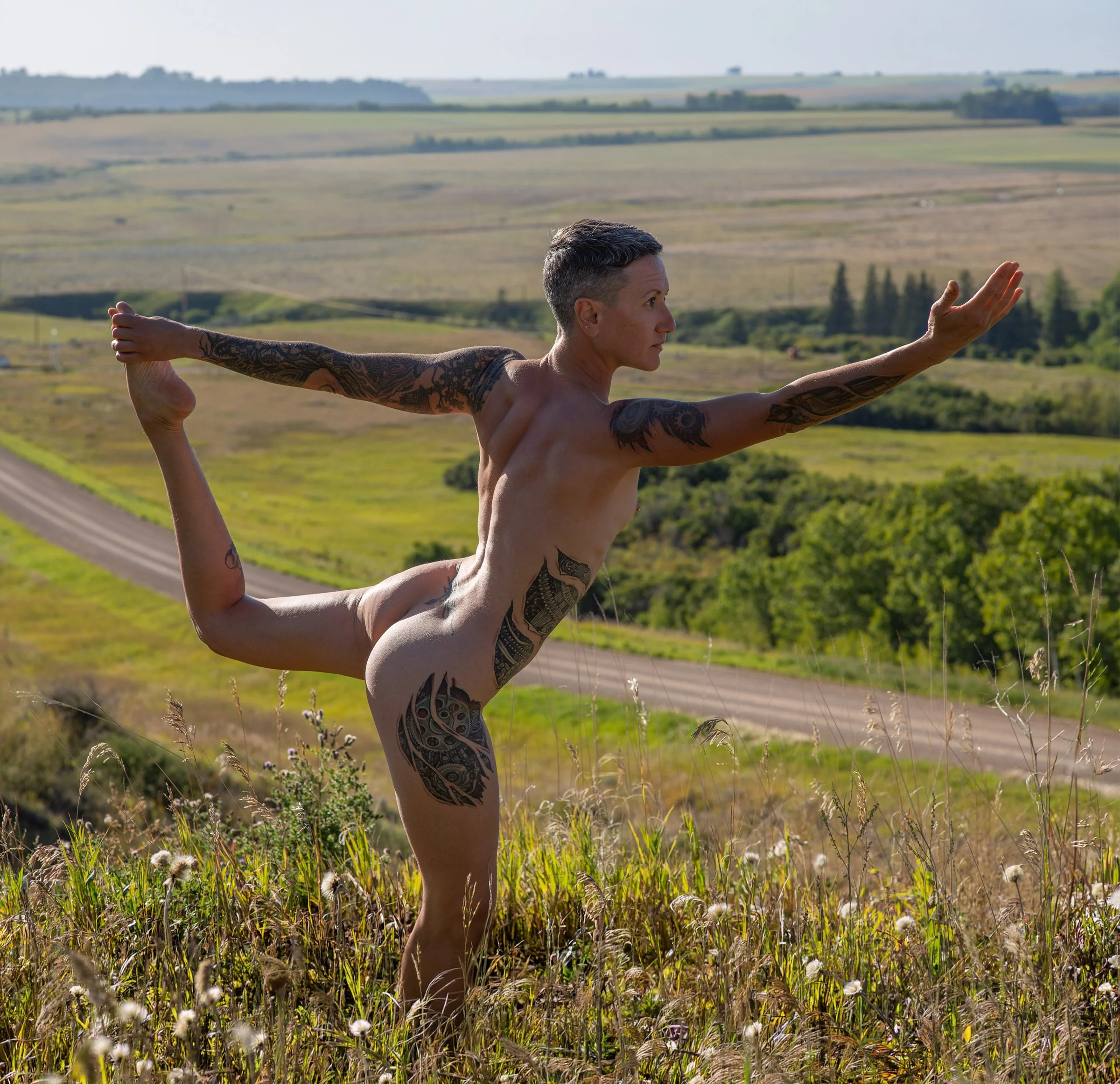 A person with short hair and tattoos practicing yoga outdoors in a grassy field with rolling hills in the background.