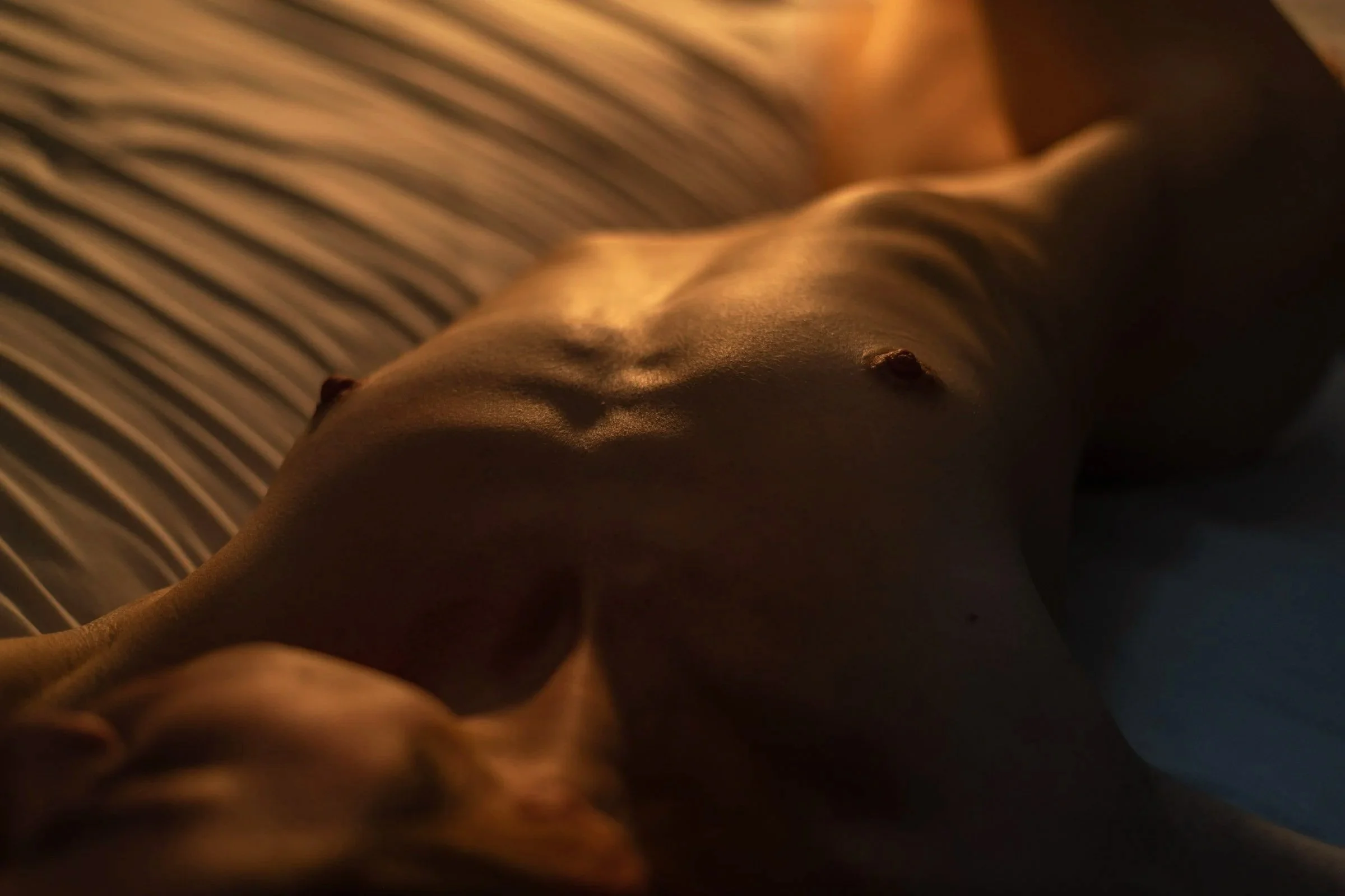Close-up of a person's torso and abdomen, with a blurred background of striped fabric, warm lighting highlighting the skin.