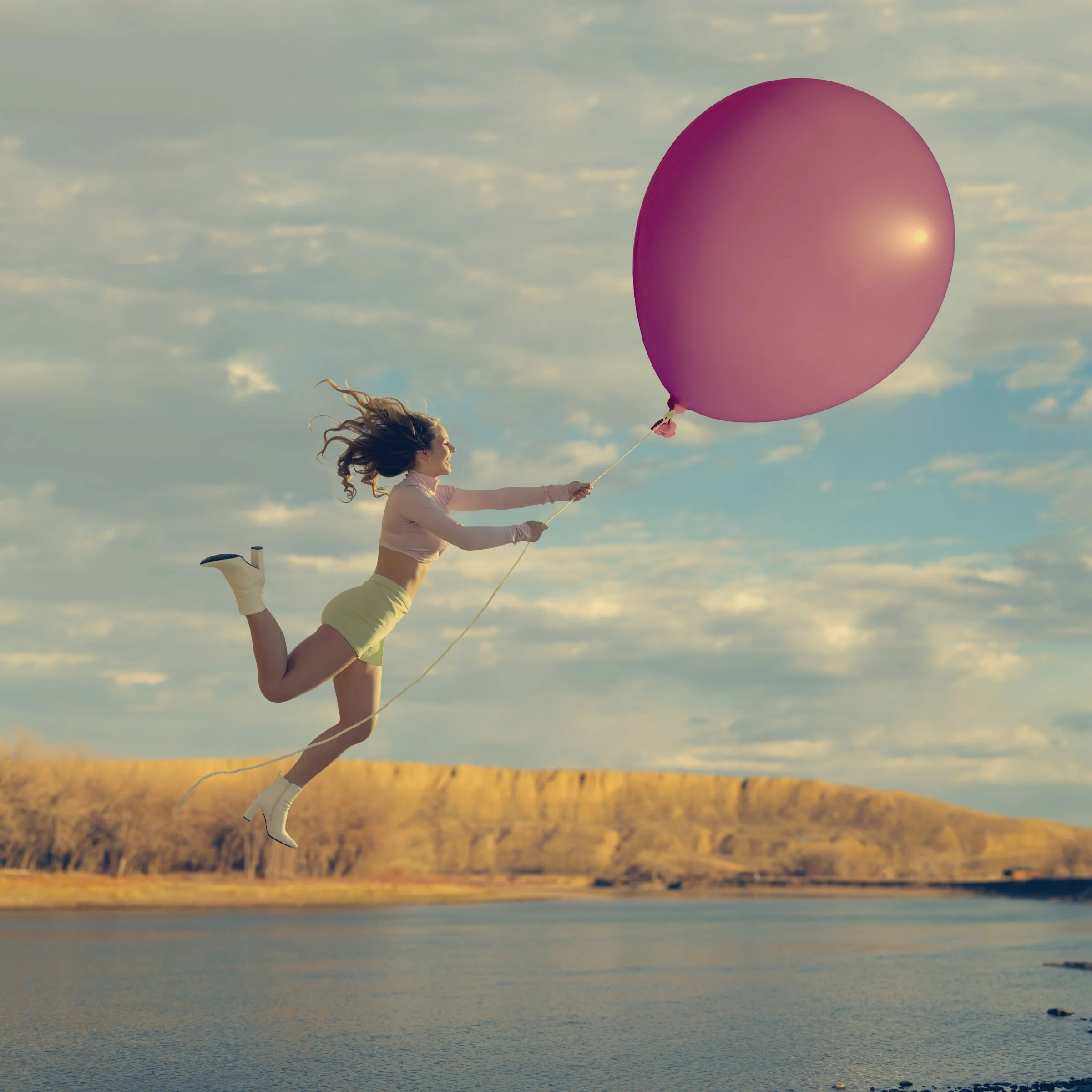 A woman with curly hair wearing white sneakers and pastel clothing, performing a yoga pose outdoors, holding a large pink balloon with a sunset and cloudy sky in the background.