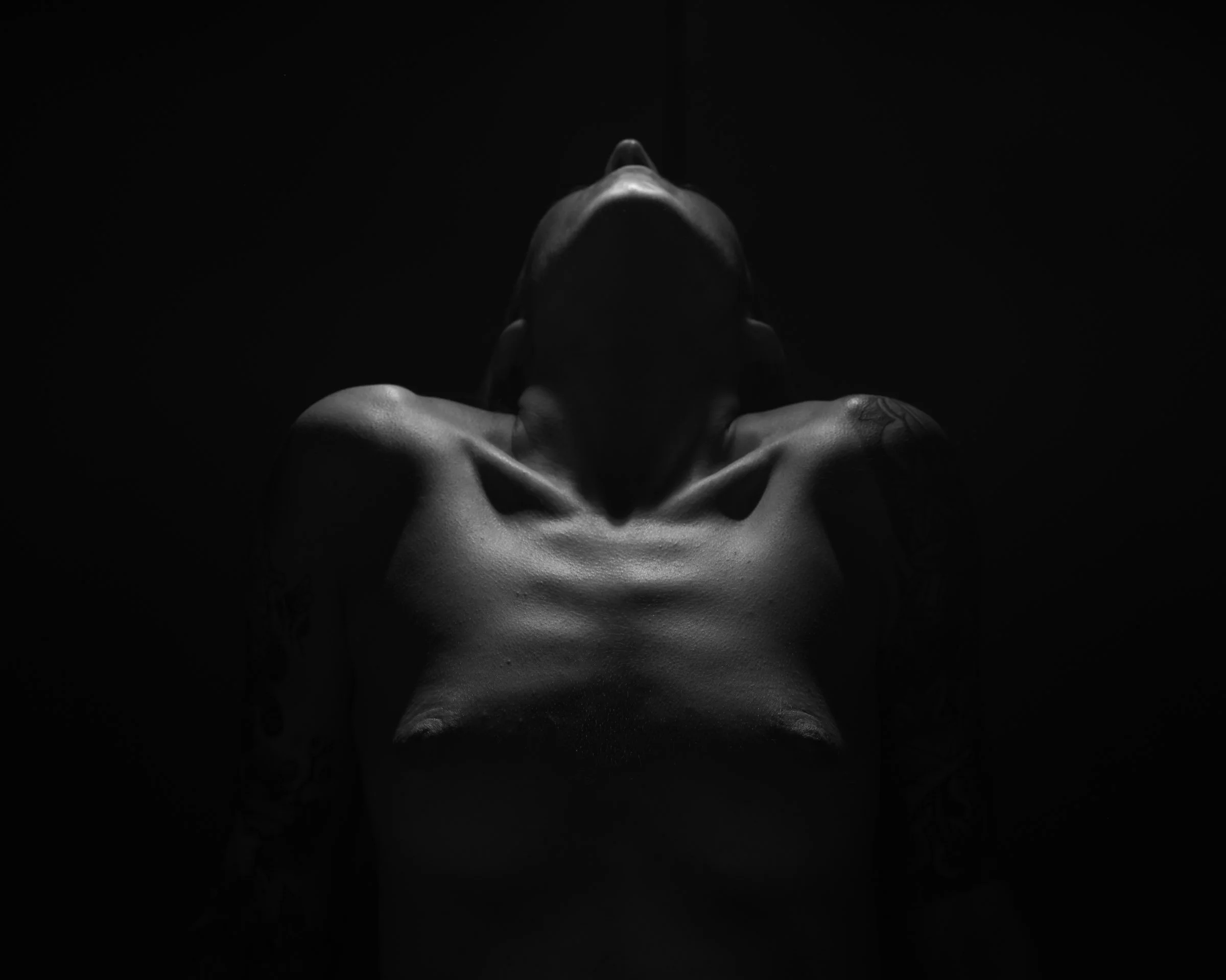 A woman with a tattoo on her wrist, topless, with her head tilted back, against a black background, illuminated by soft lighting.
