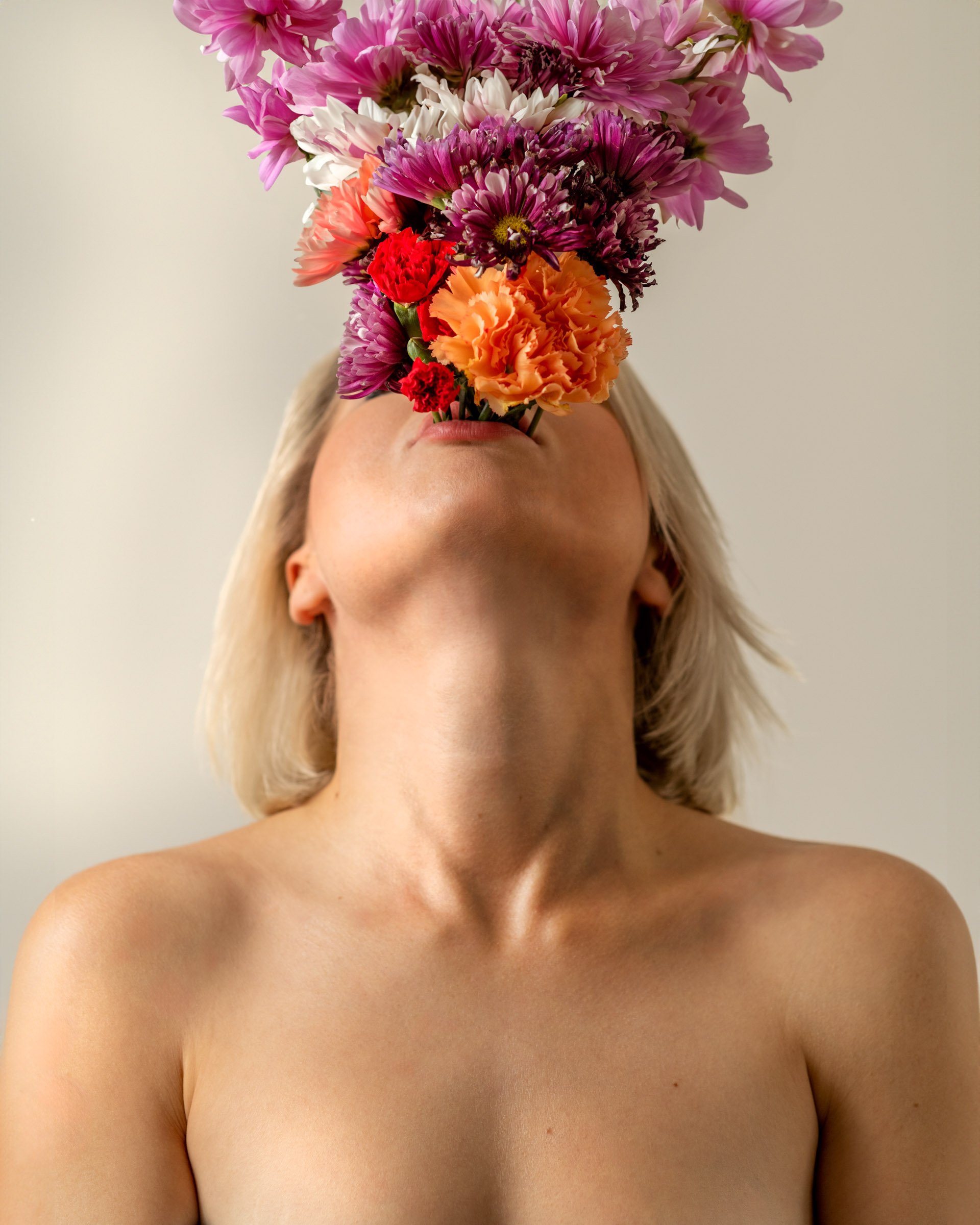 A person with blonde hair, shown from the chin up, with a large bouquet of pink, purple, white, red, and orange flowers covering their face, against a neutral background.