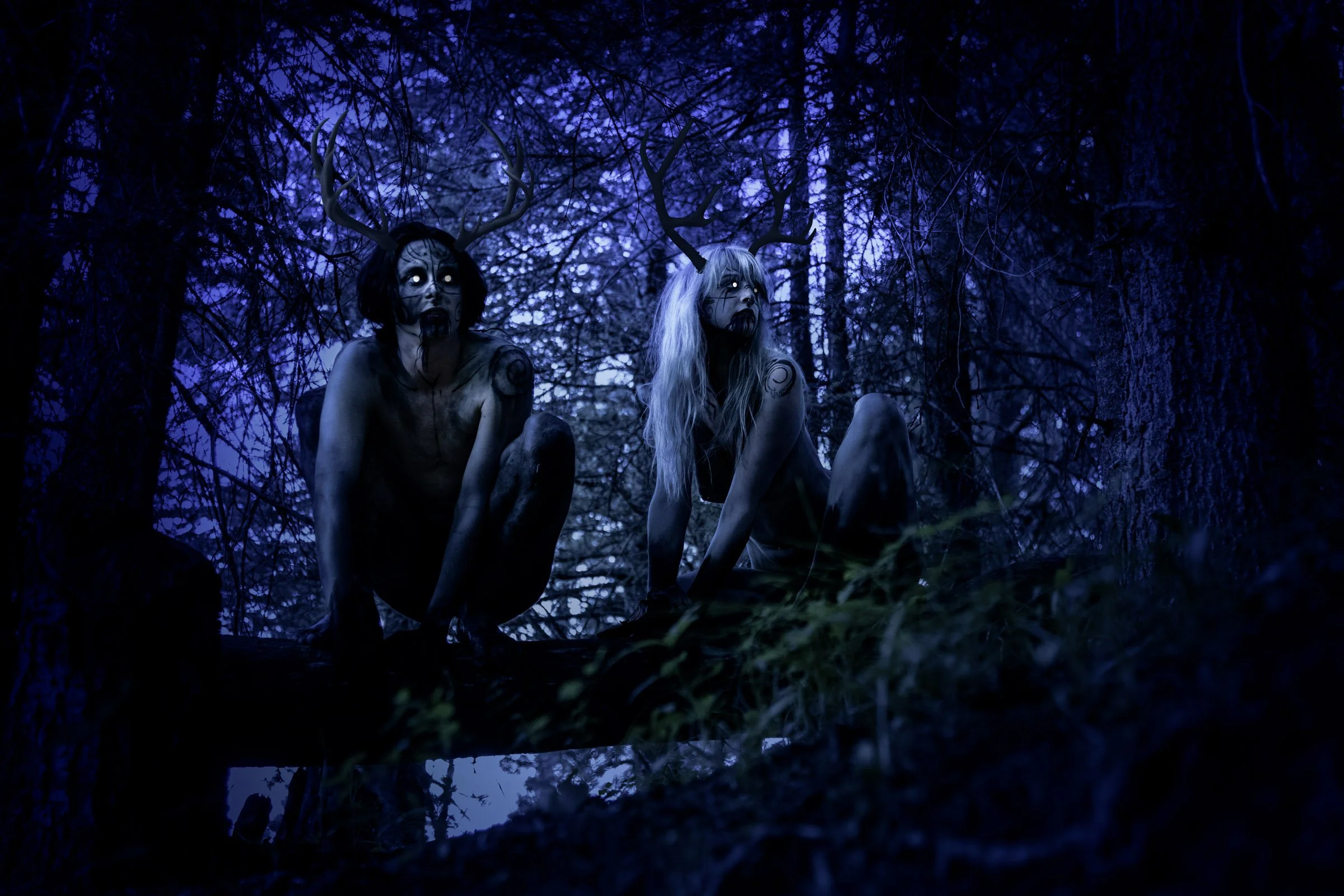 Two humanoid figures with antlers and dark makeup sitting in a dark, eerie forest at night.
