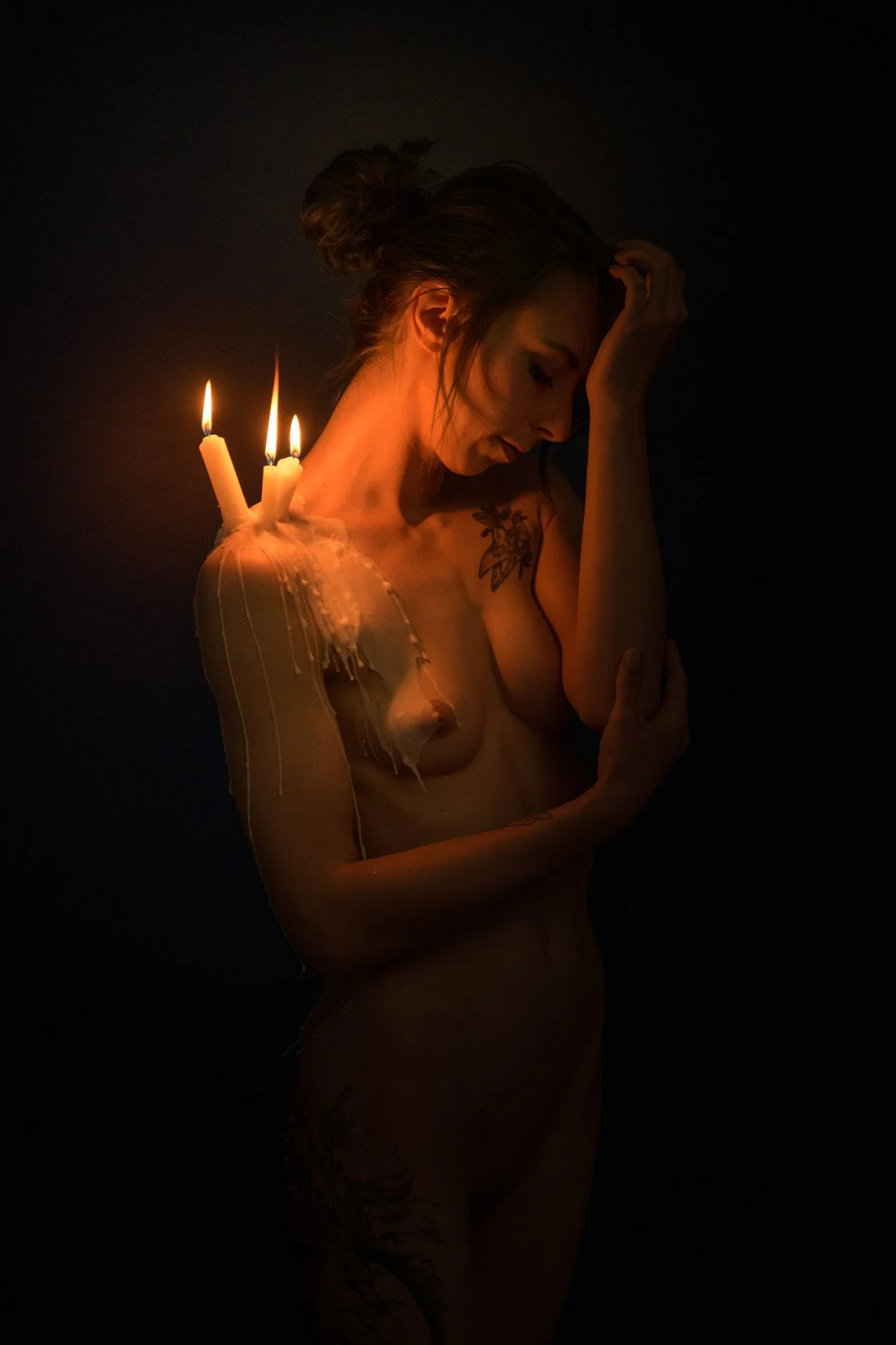 A woman with tattoos is holding her head with one hand, with three lit candles burning on her shoulder, and is surrounded by darkness.