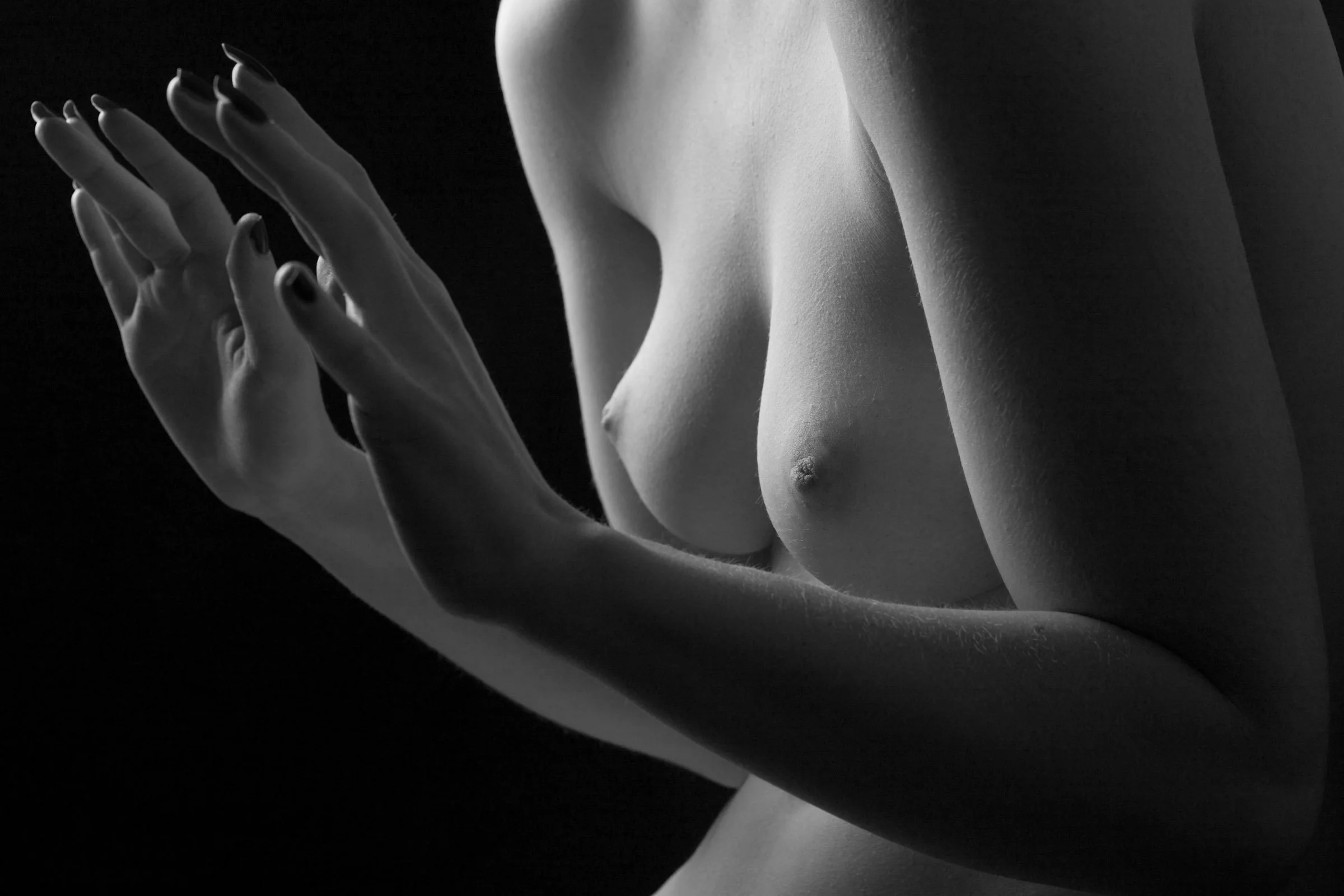 Close-up of a woman in black and white, showing her bare shoulder, part of her torso, and hands with fingers touching near her face, emphasizing the contours of her body and skin texture.