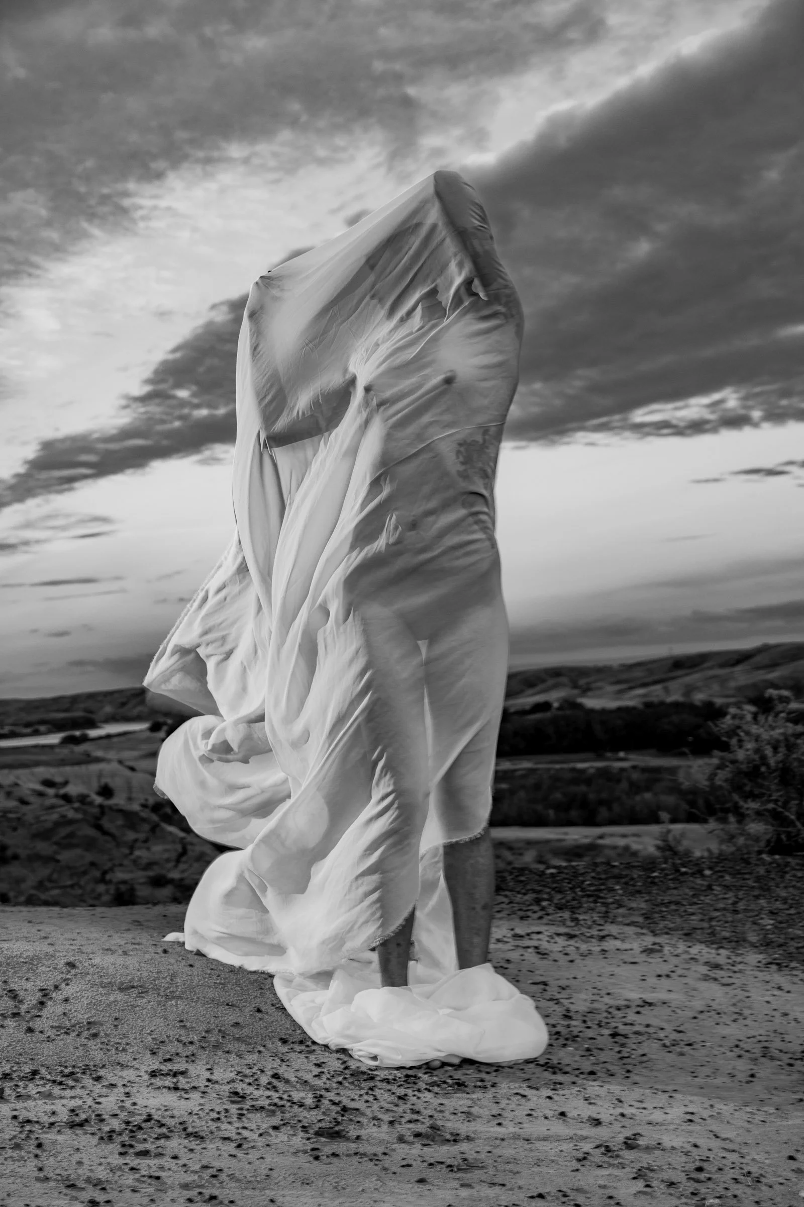A person standing on a beach with a flowing white dress or fabric, with an overcast sky and clouds in the background.