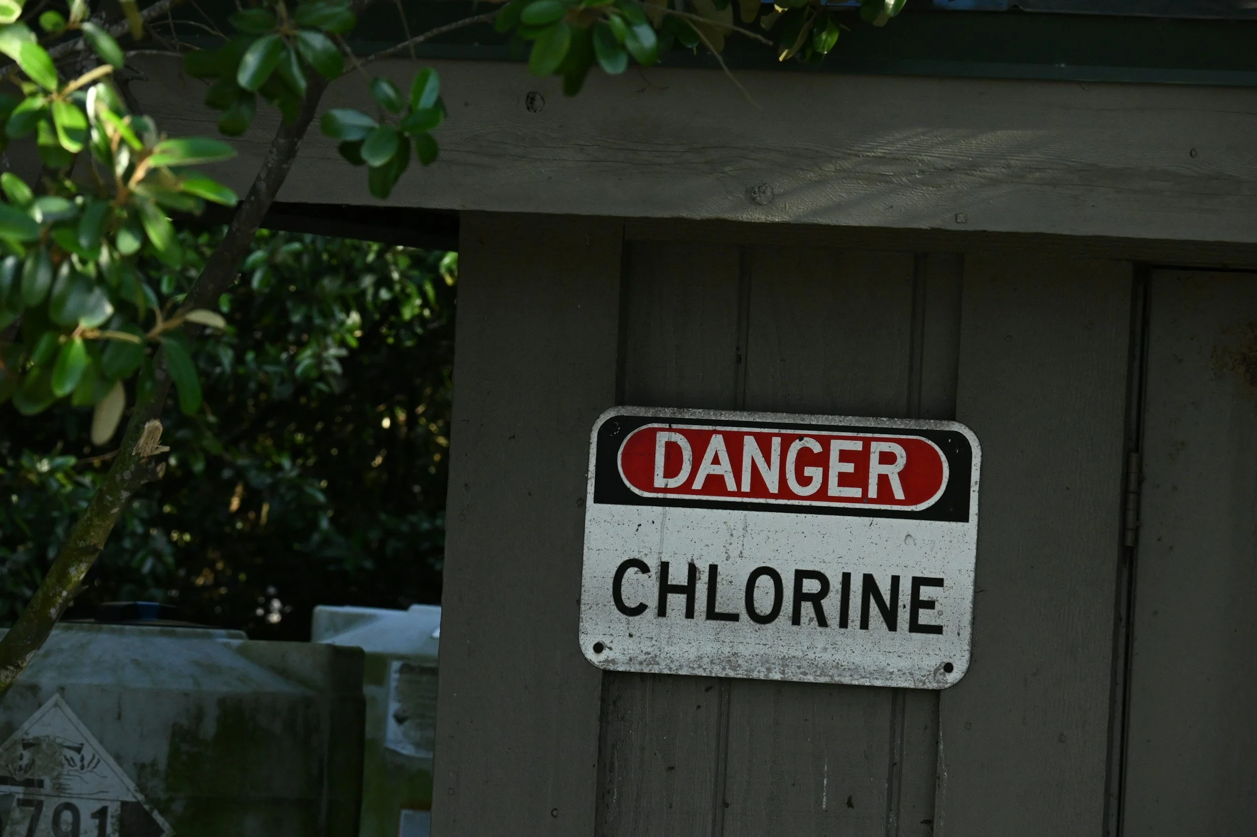 A warning sign on a gray wooden structure that reads 'DANGER CHLORINE'. Green leaves and branches are partially visible on the left side of the image.