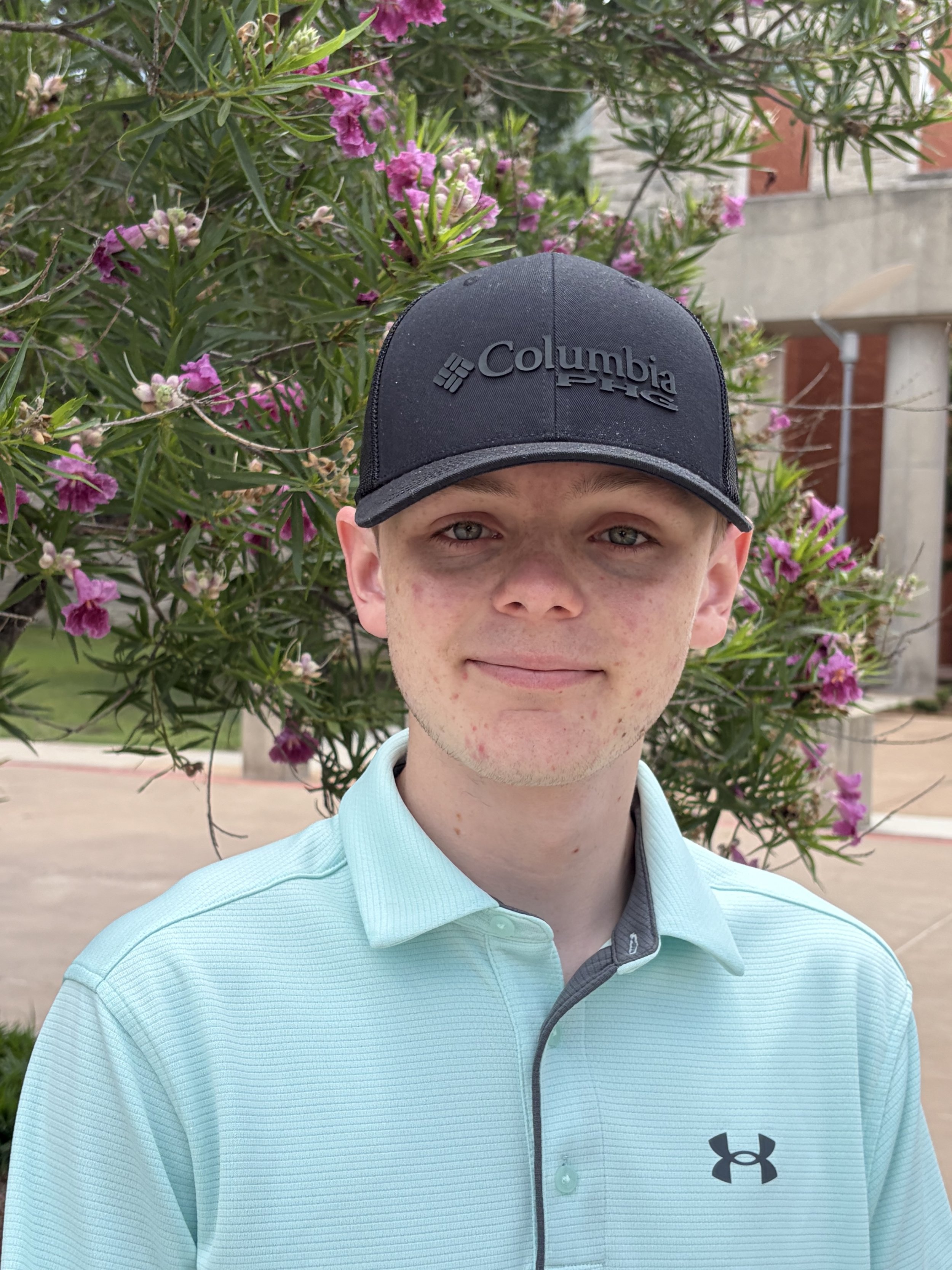 A young man with light skin, light green eyes, and freckles, wearing a black Columbia cap and a light blue Under Armour polo shirt, stands in front of pink flowering bushes outdoors.