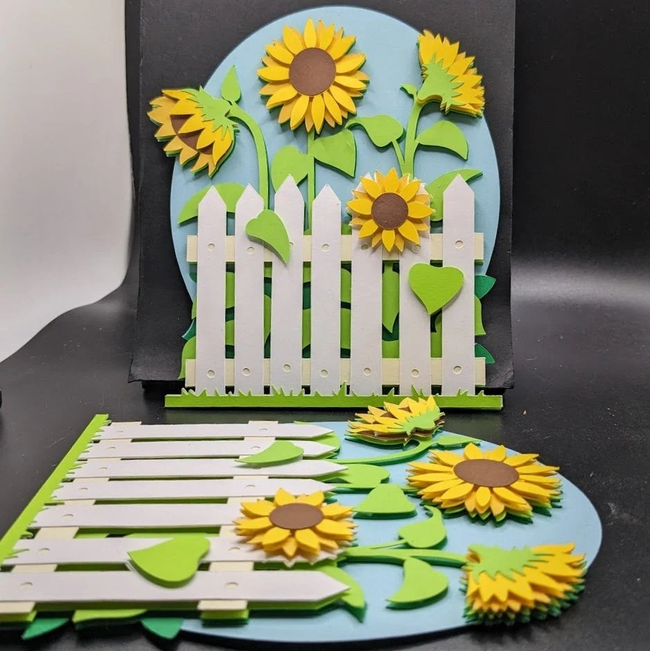 Stacked and assembled paper craft of sunflowers, a white picket fence, green leaves, and a blue oval background.