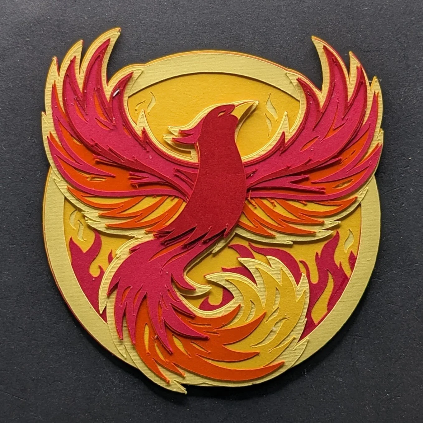 A layered paper craft of a phoenix with red and orange flames, set against a yellow circular background.